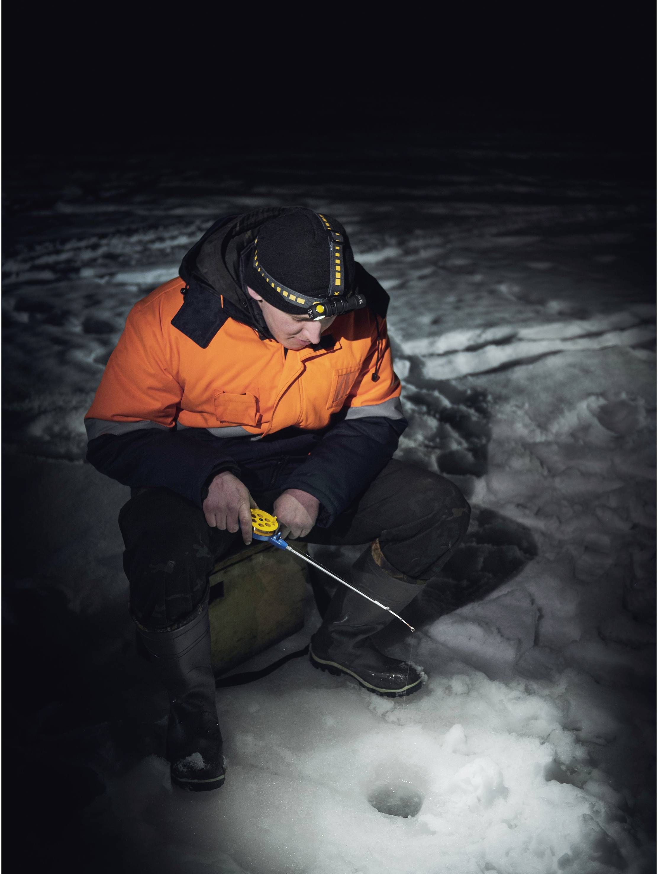 Une personne portant une veste orange vif est assise sur un lac gelé, pêchant sur glace à travers un trou dans la glace. Environnement nocturne sombre.