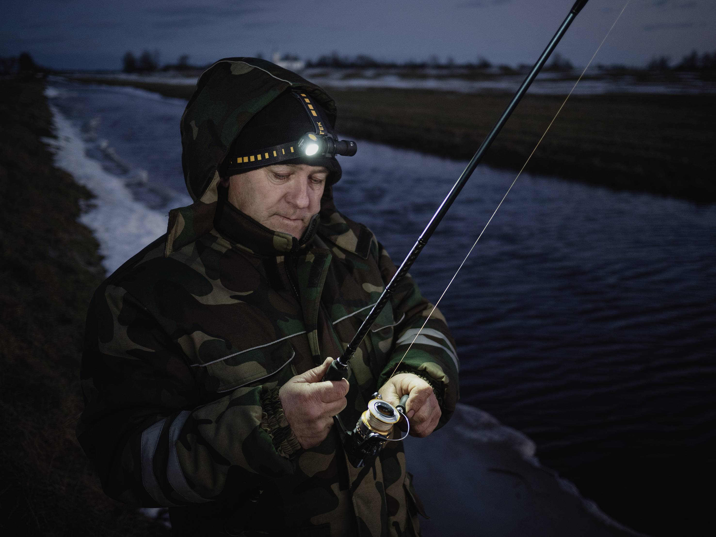 Une personne en tenue de camouflage pêche la nuit au bord d'une rivière. Elle porte une lampe frontale qui éclaire son visage et sa canne à pêche.