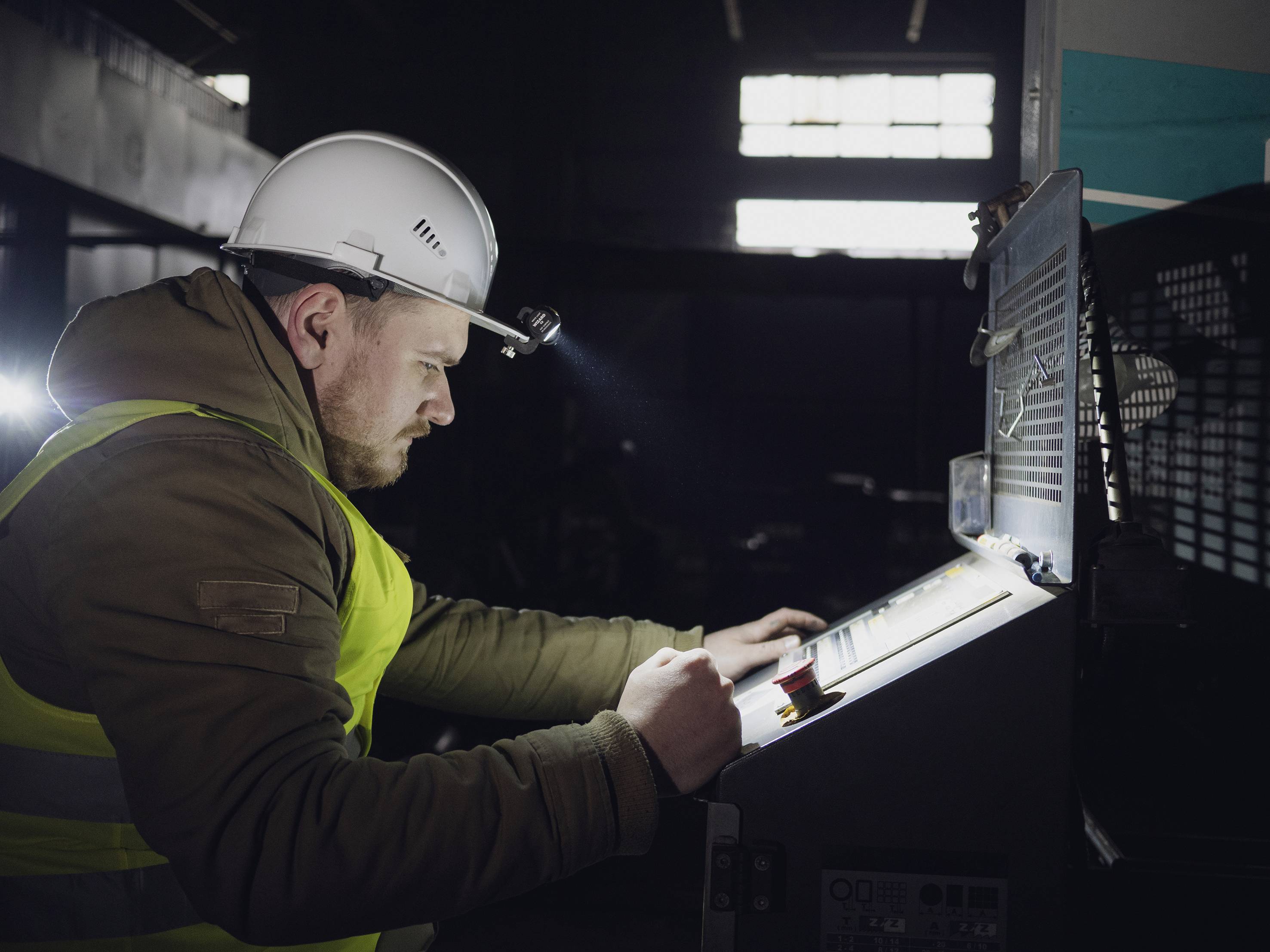 Un travailleur portant un casque et un gilet de protection actionne une machine industrielle avec un écran de contrôle dans un environnement sombre.