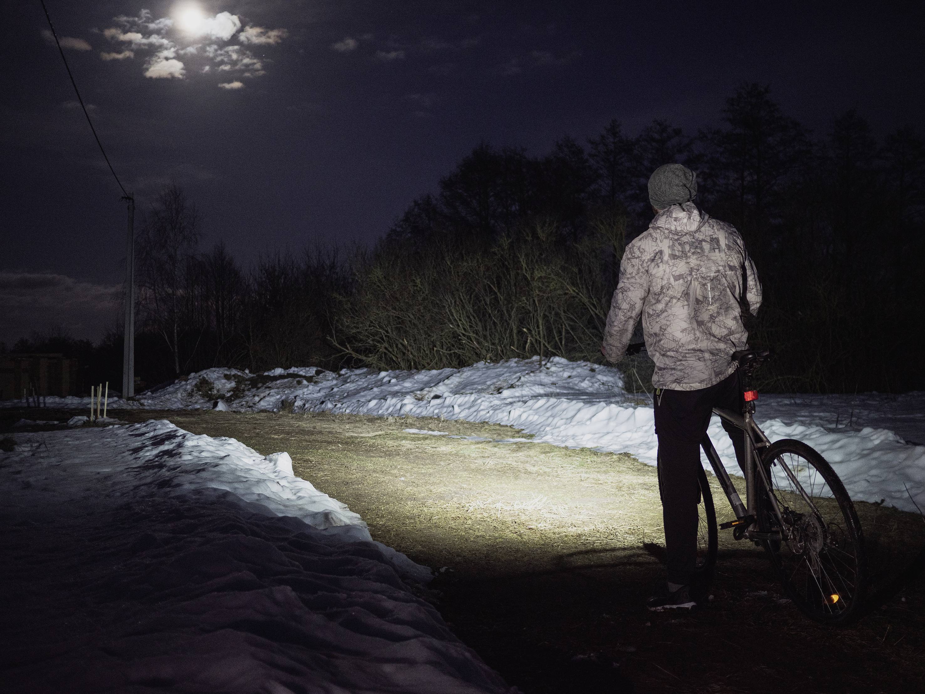 Une personne circule à vélo sur un chemin enneigé pendant la nuit. Le chemin est éclairé par la lumière du phare du vélo, et la lune est visible.