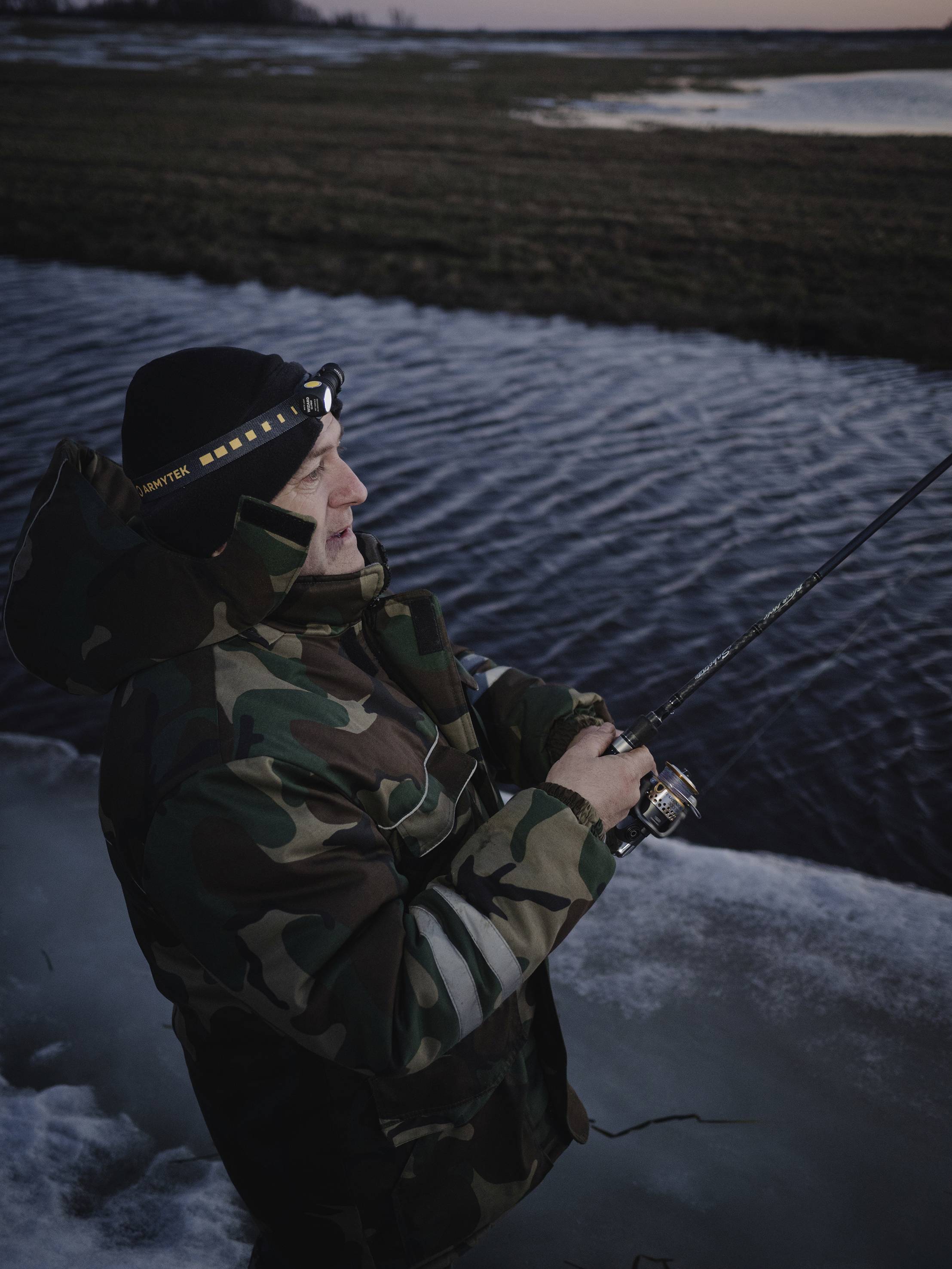 Une personne en tenue de camouflage pêche avec une lampe frontale sur la berge d'une rivière au crépuscule.