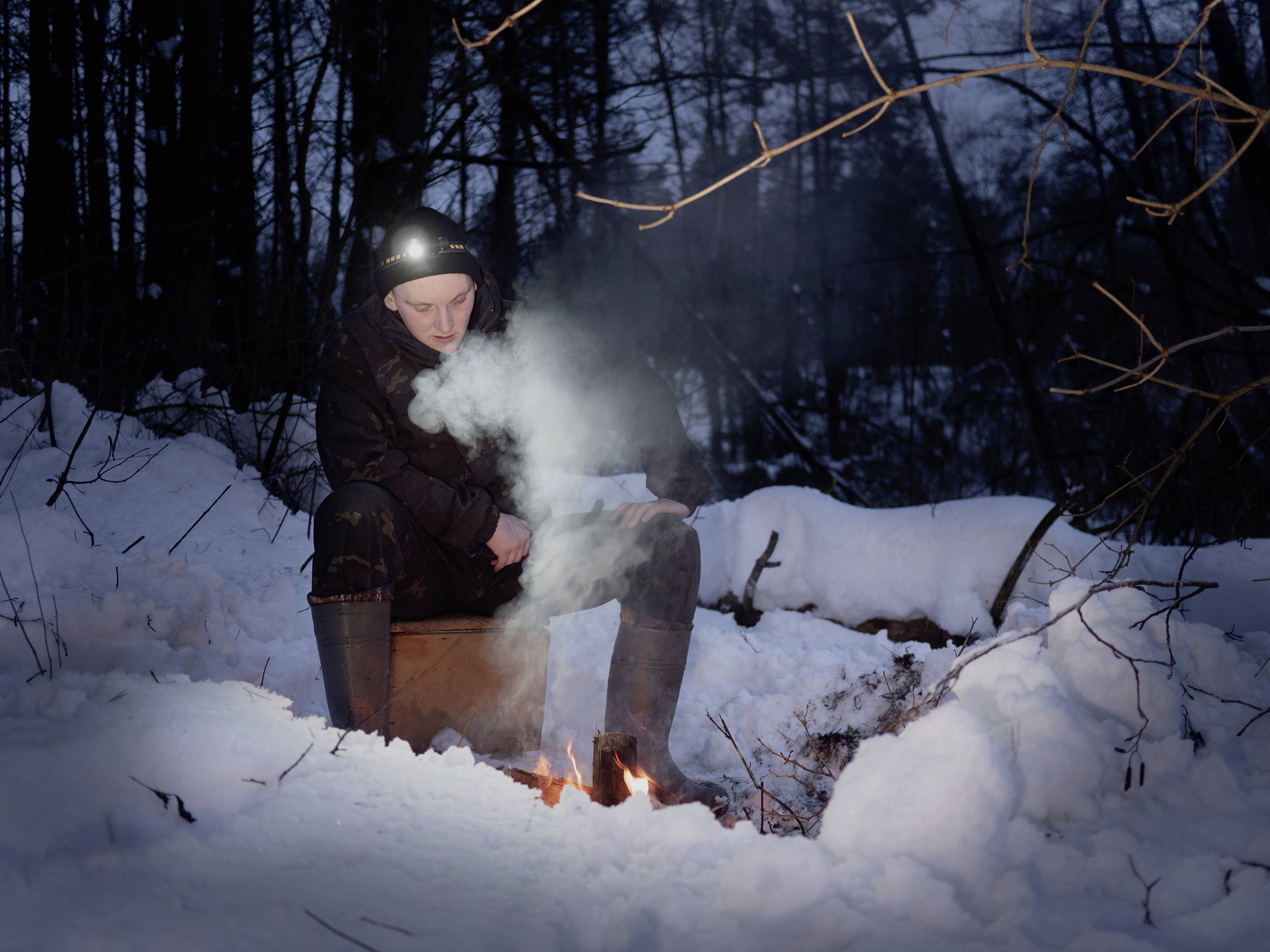 Une personne est assise dans la forêt enneigée, la nuit, près d'un petit feu, portant une lampe frontale. L'ambiance est calme et atmosphérique.
