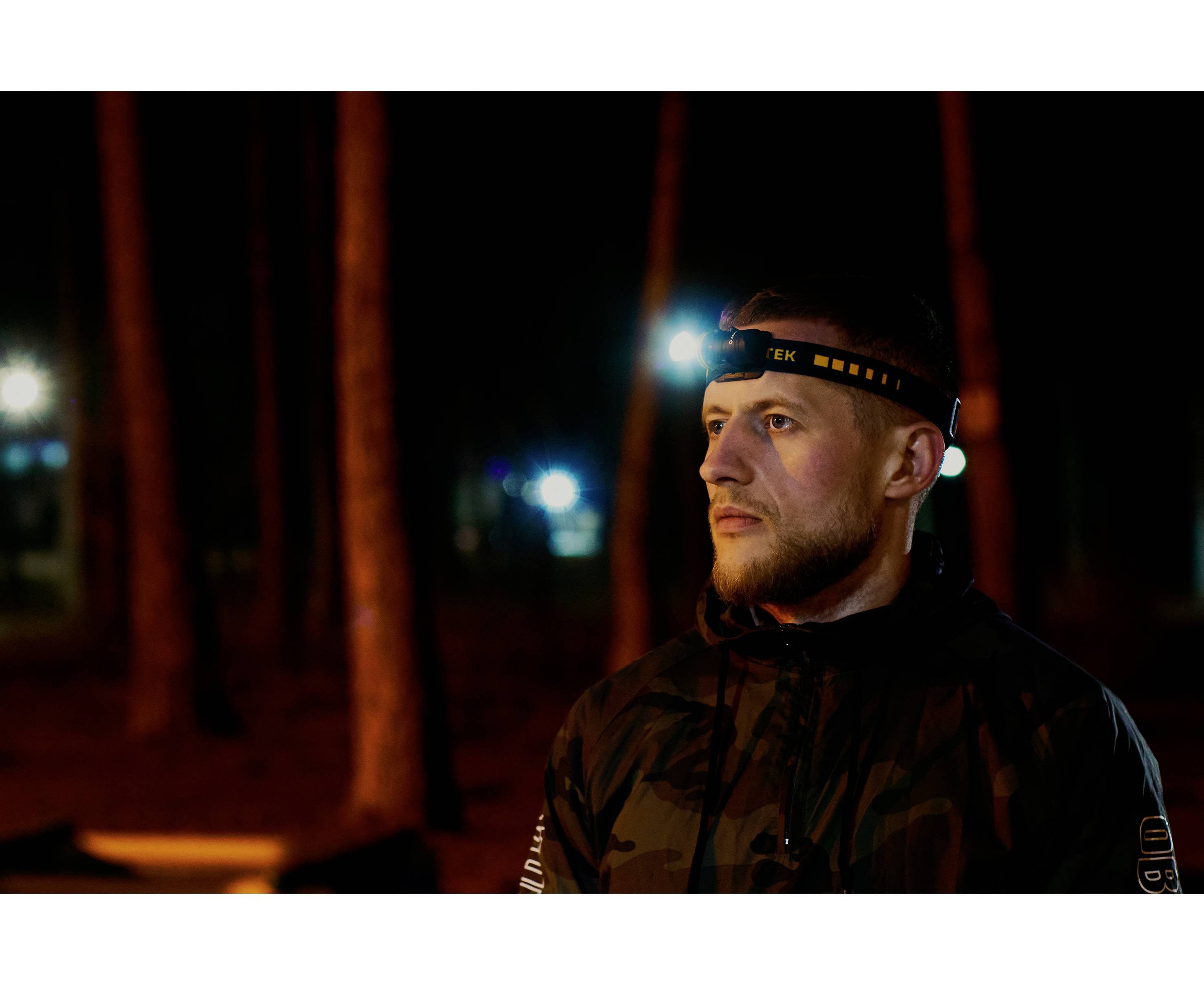 Un homme portant une lampe frontale se tient dans la forêt le soir. Il porte une veste et regarde sérieusement au loin. Des arbres sont visibles en arrière-plan.