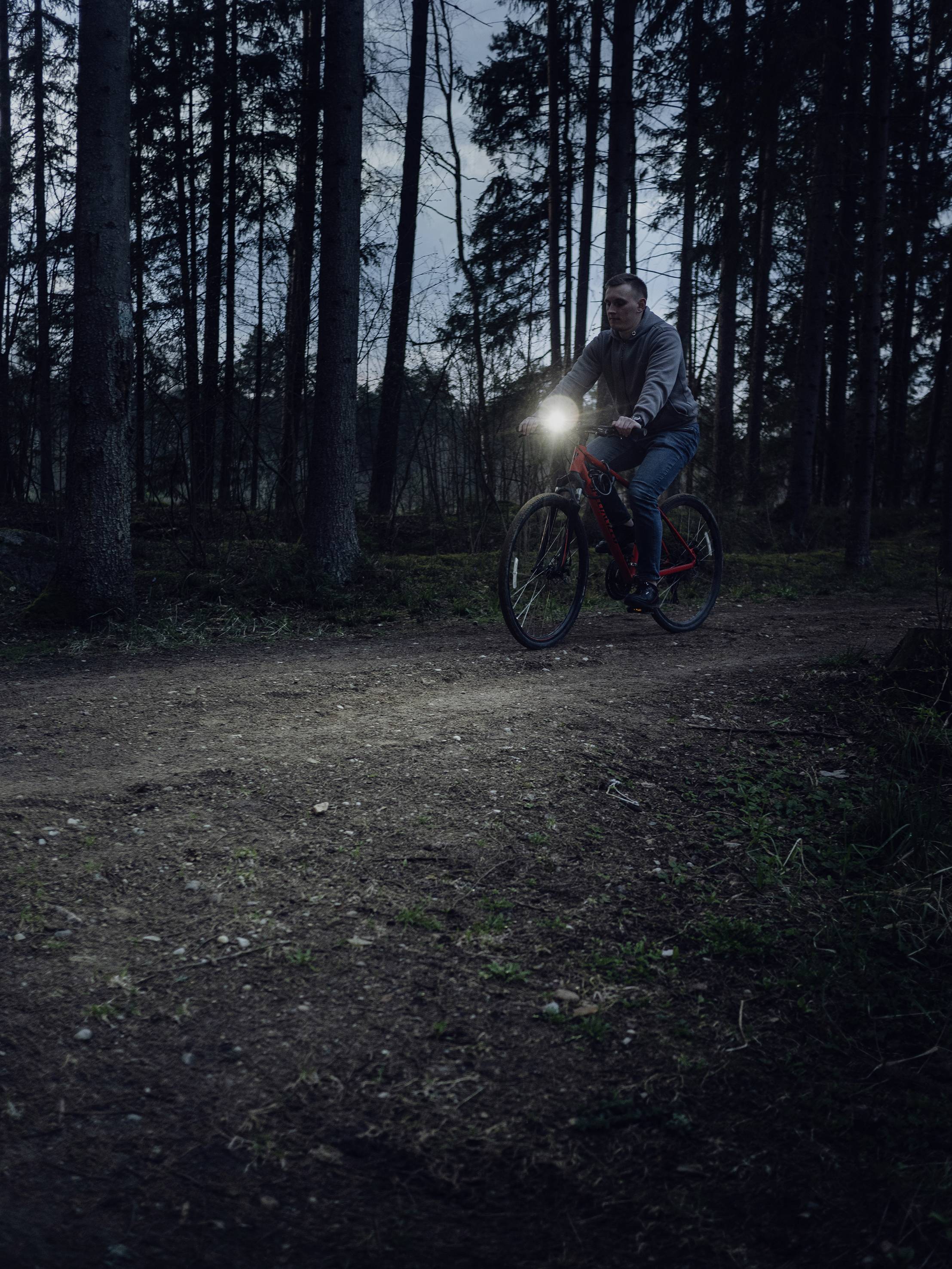 Une personne fait du vélo de nuit sur un chemin forestier, éclairée par sa lampe, entourée d'arbres imposants.