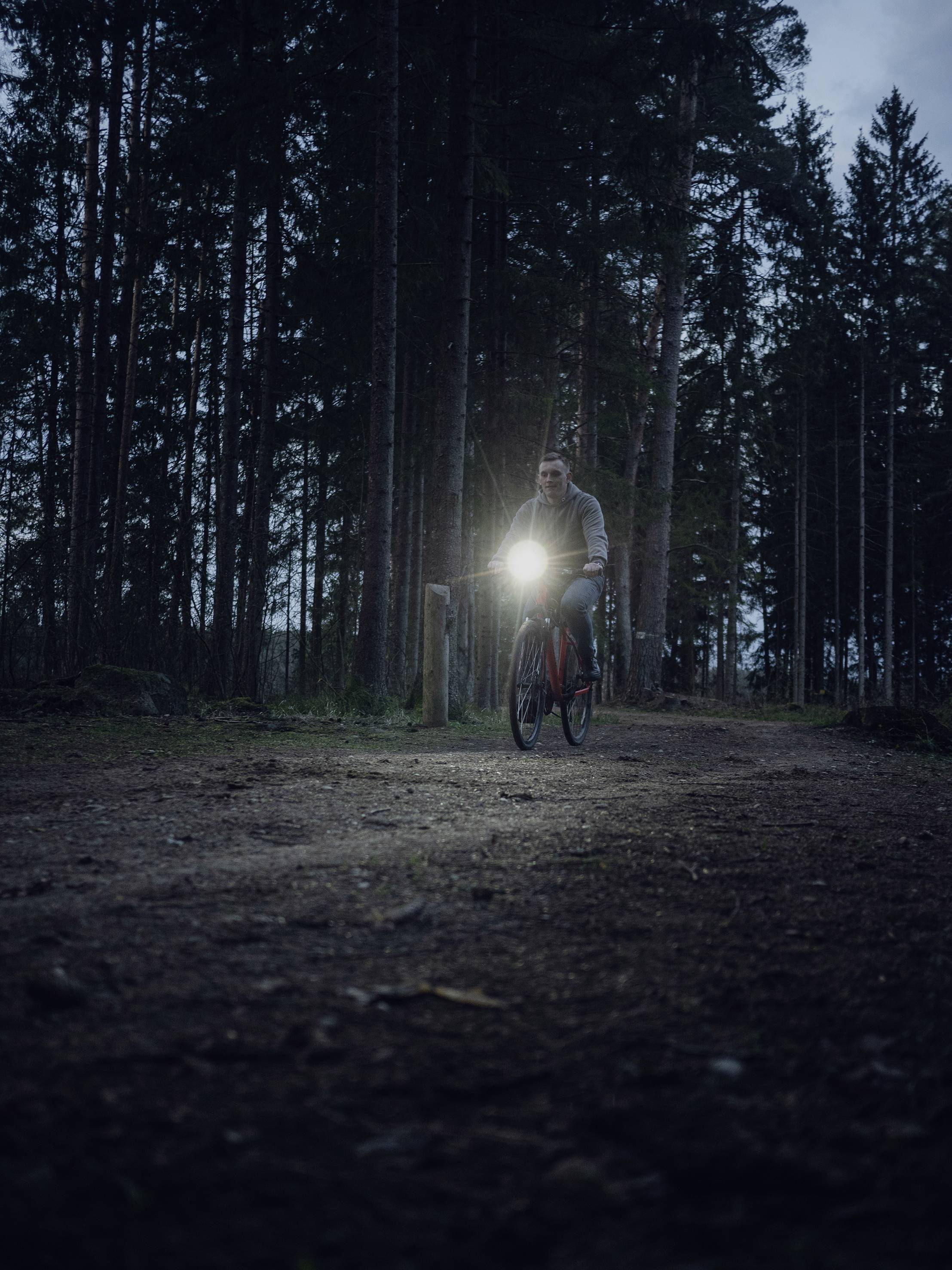 Une personne pédale à vélo sur un chemin forestier au crépuscule. Le phare du vélo éclaire le chemin.