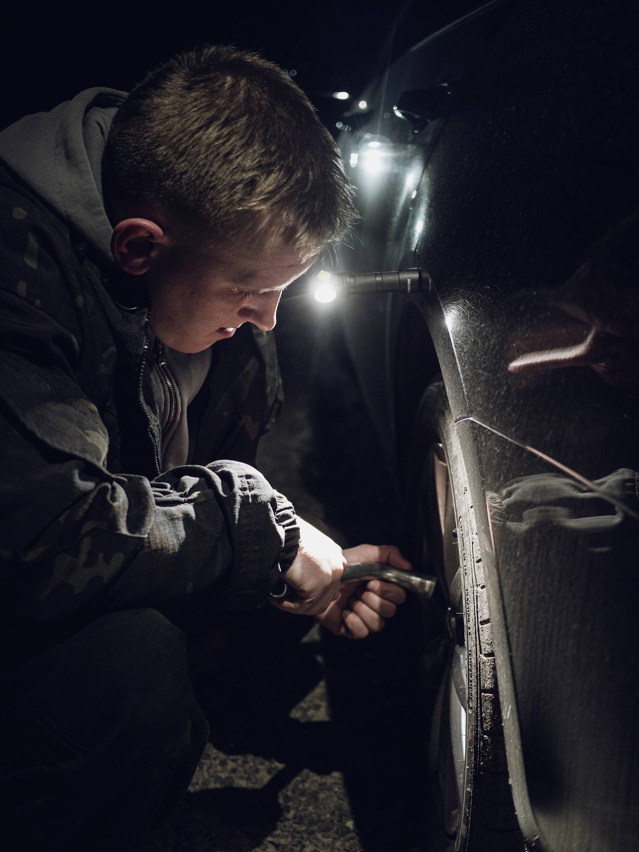 Un homme change un pneu la nuit sur une route sombre. Une lampe de poche éclaire son travail.