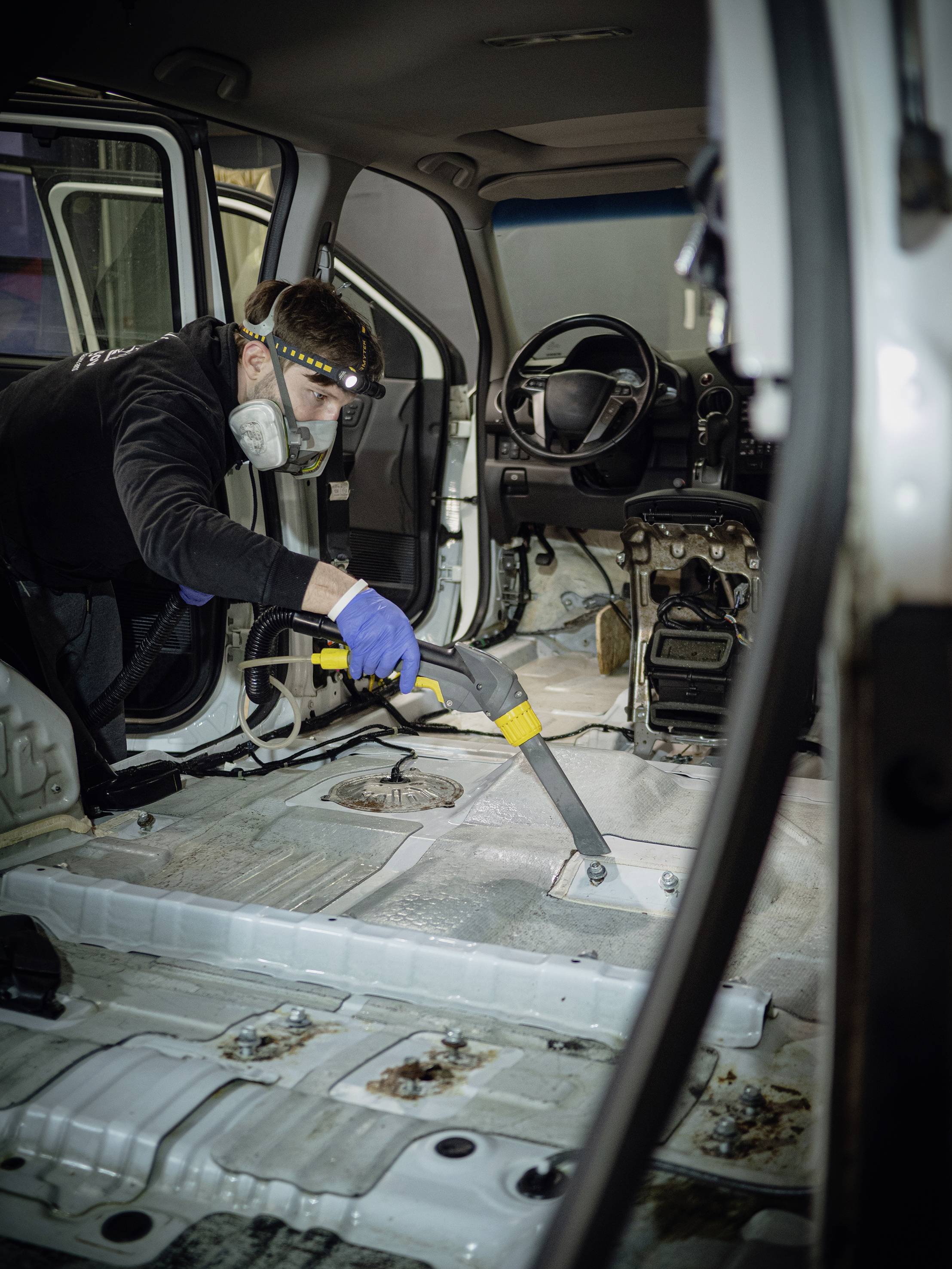 Une personne nettoie l'intérieur d'une voiture sans sièges à l'aide d'un aspirateur. Un masque de protection et des gants sont portés.