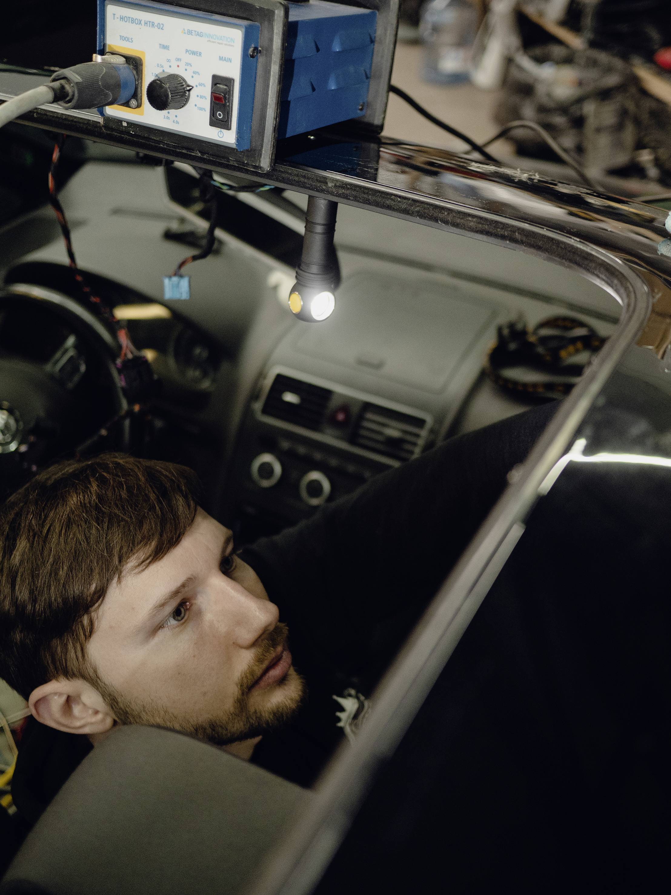 Un homme travaille sur l'électricité d'une voiture, observé par un petit point lumineux sur le pare-brise.