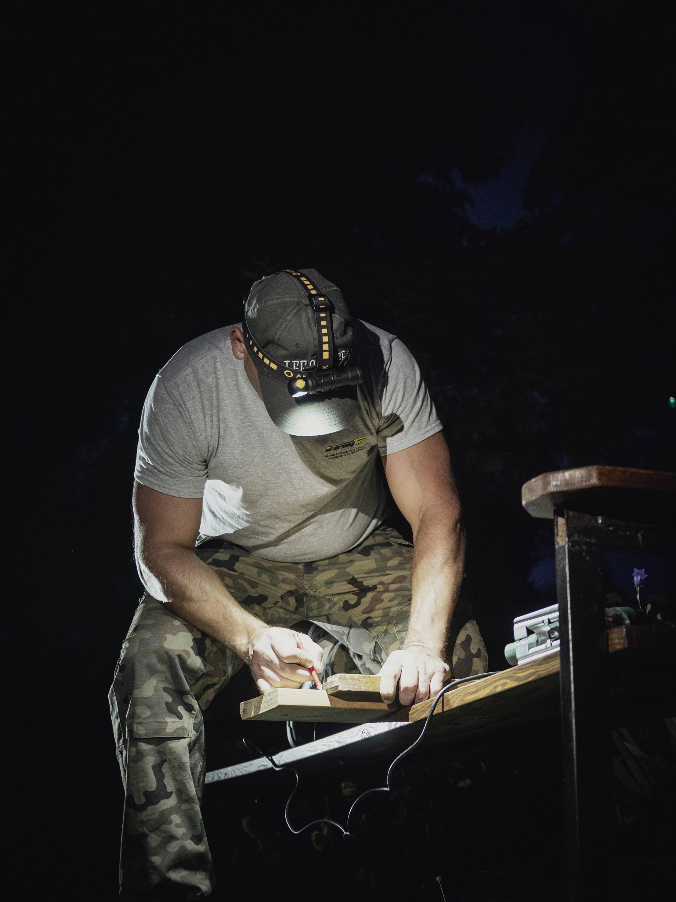 Un homme en pantalon de camouflage et t-shirt gris travaille dans l'obscurité avec un outil sur une table. Une lampe frontale éclaire sa zone de travail.