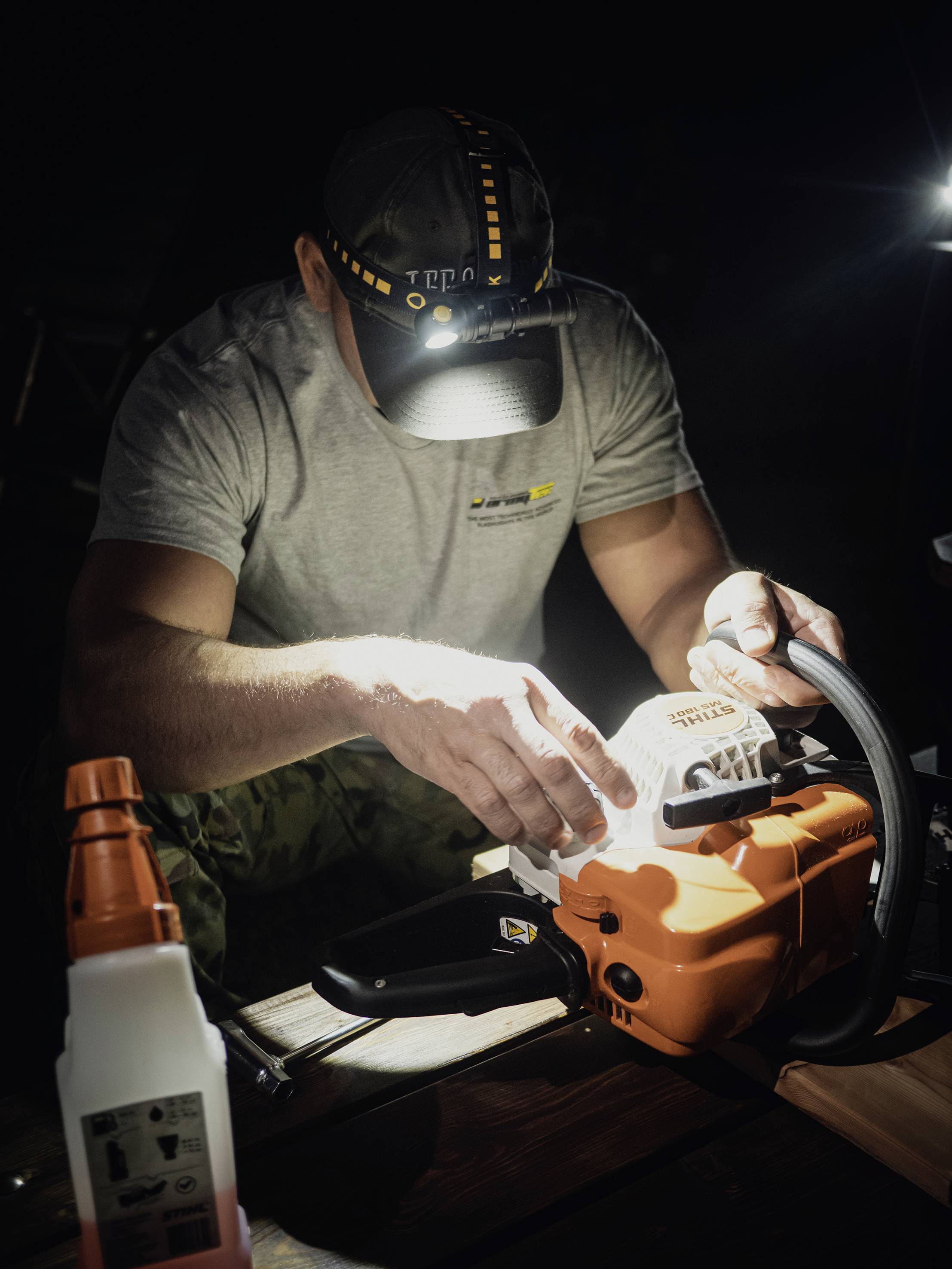 Un homme portant une lampe frontale travaille la nuit sur une tronçonneuse posée sur une table et utilise une lime pour entretenir la chaîne.
