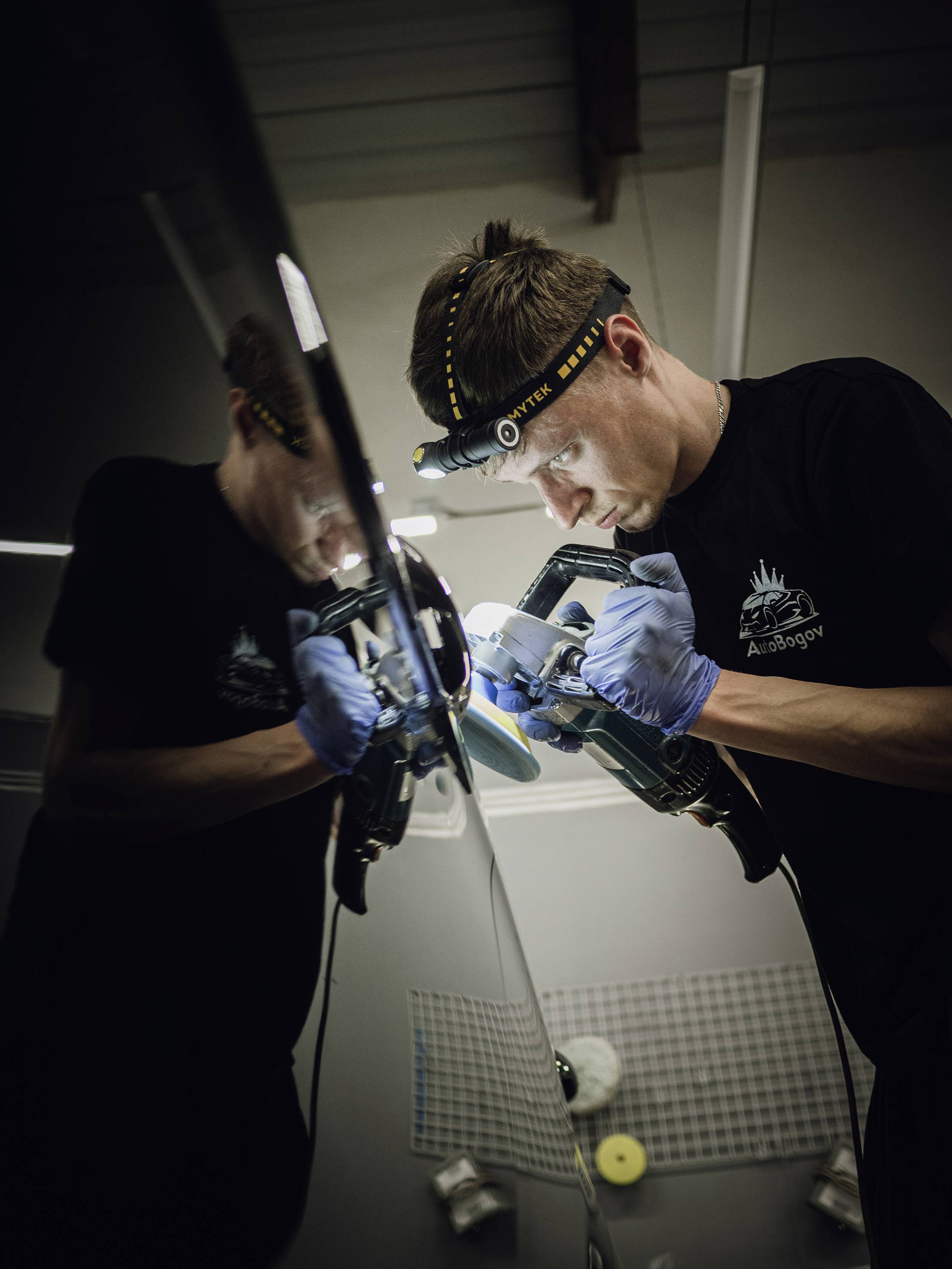 Un homme portant une lampe frontale polit soigneusement la surface d'un véhicule. Il porte des gants et travaille dans un environnement d'atelier.