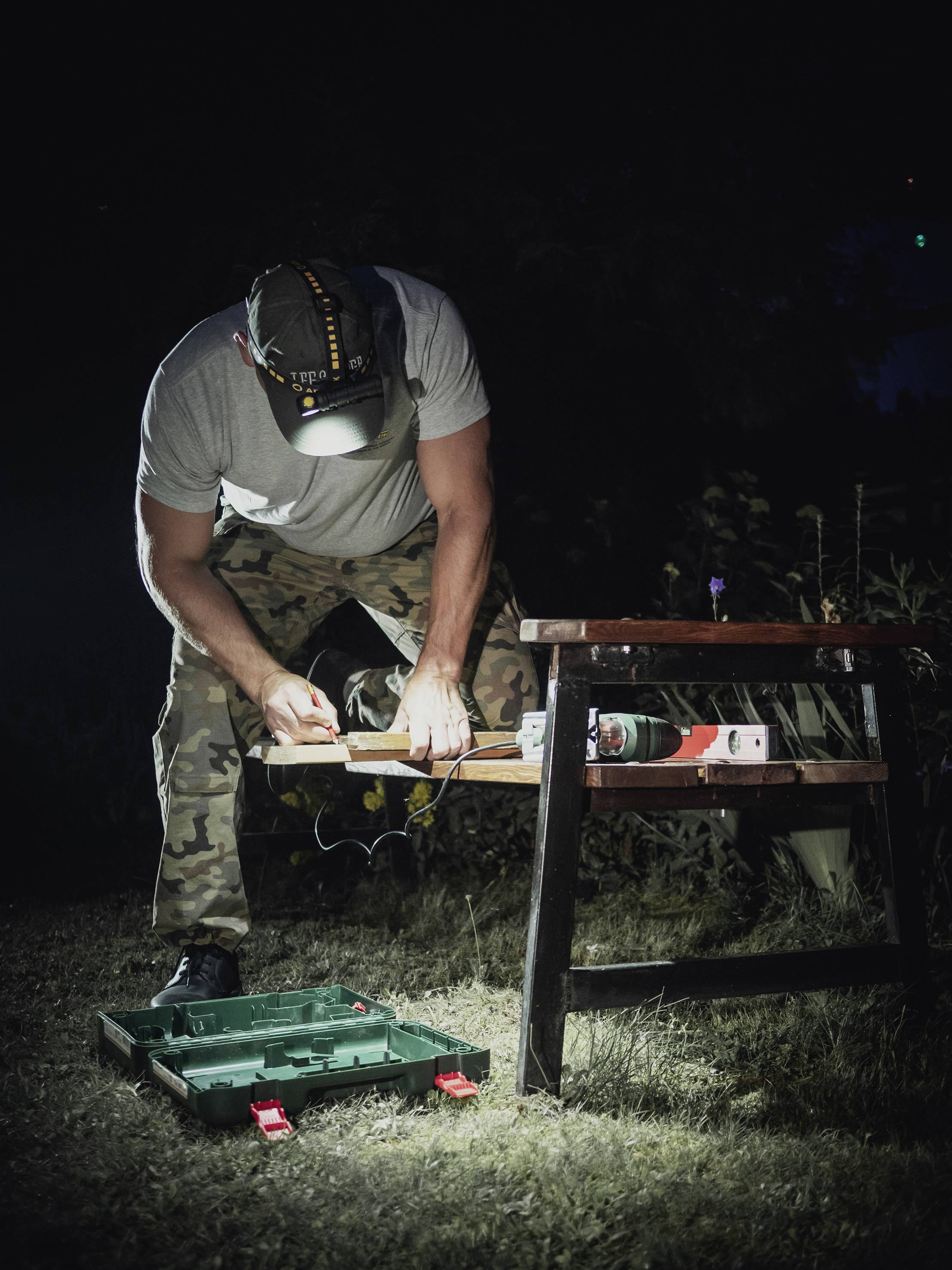 Un homme portant un pantalon de camouflage travaille dehors la nuit avec une boîte à outils ; il éclaire son travail à l'aide d'une lampe frontale.