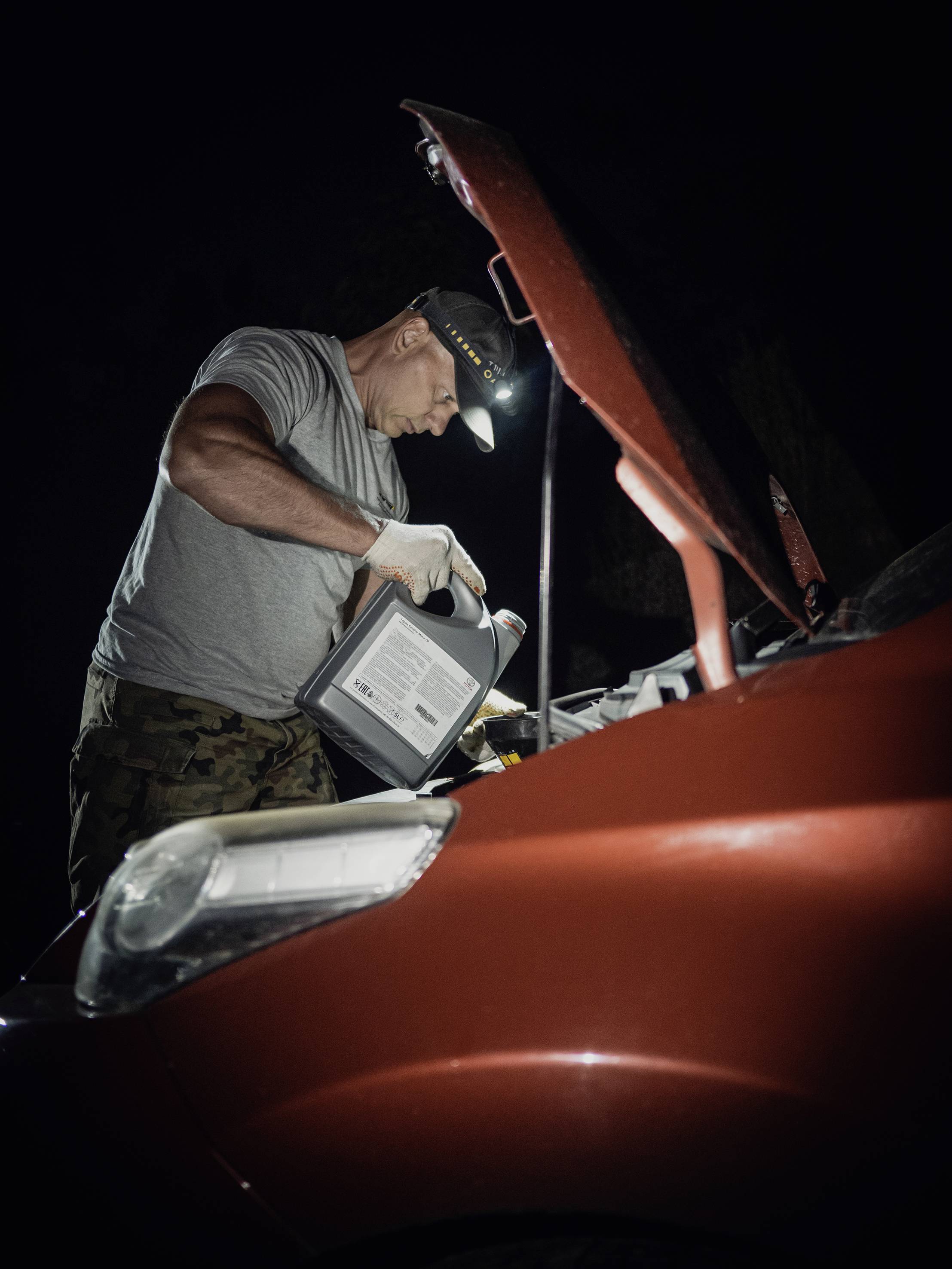 Une personne en tenue de travail ajoute de l'huile dans le moteur d'une voiture rouge. La scène se déroule la nuit, éclairée par une lampe frontale.
