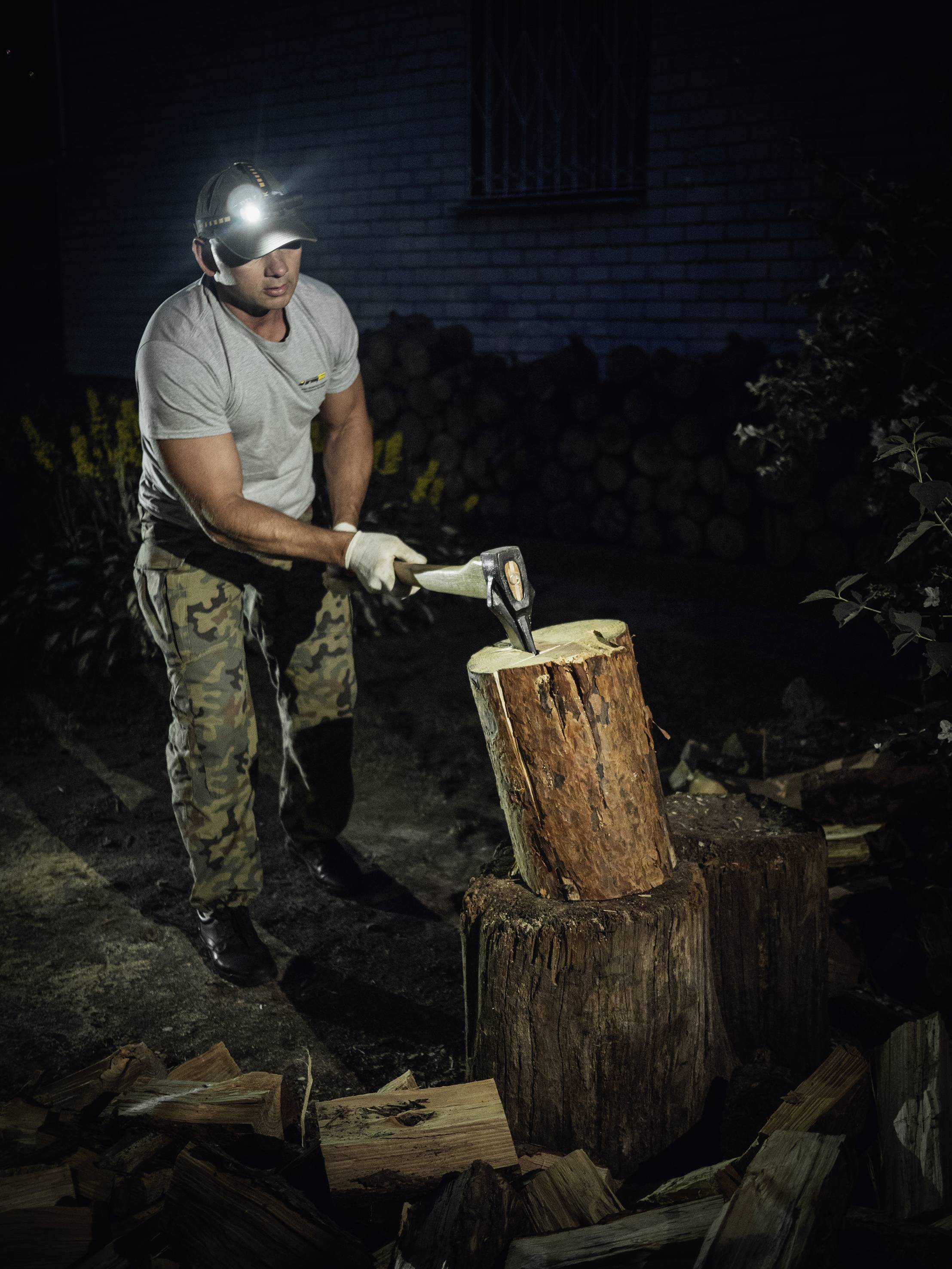 Une personne fend du bois la nuit à la hache. Elle porte une lampe frontale et un pantalon de camouflage. Des piles de bois sont visibles en arrière-plan.