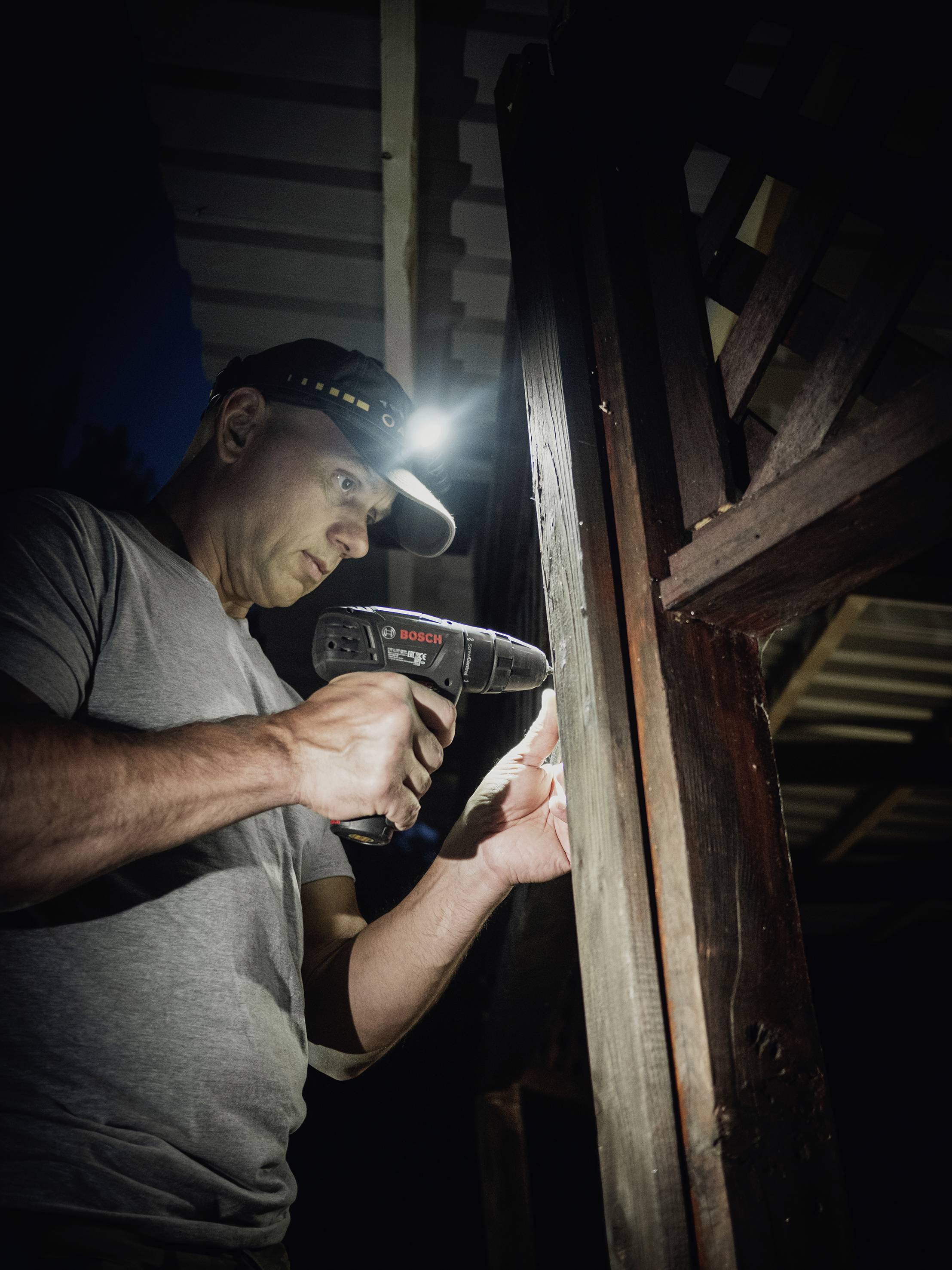 Un homme travaille le soir à l'extérieur avec une perceuse sur une poutre en bois, une lampe frontale éclairant sa zone de travail.