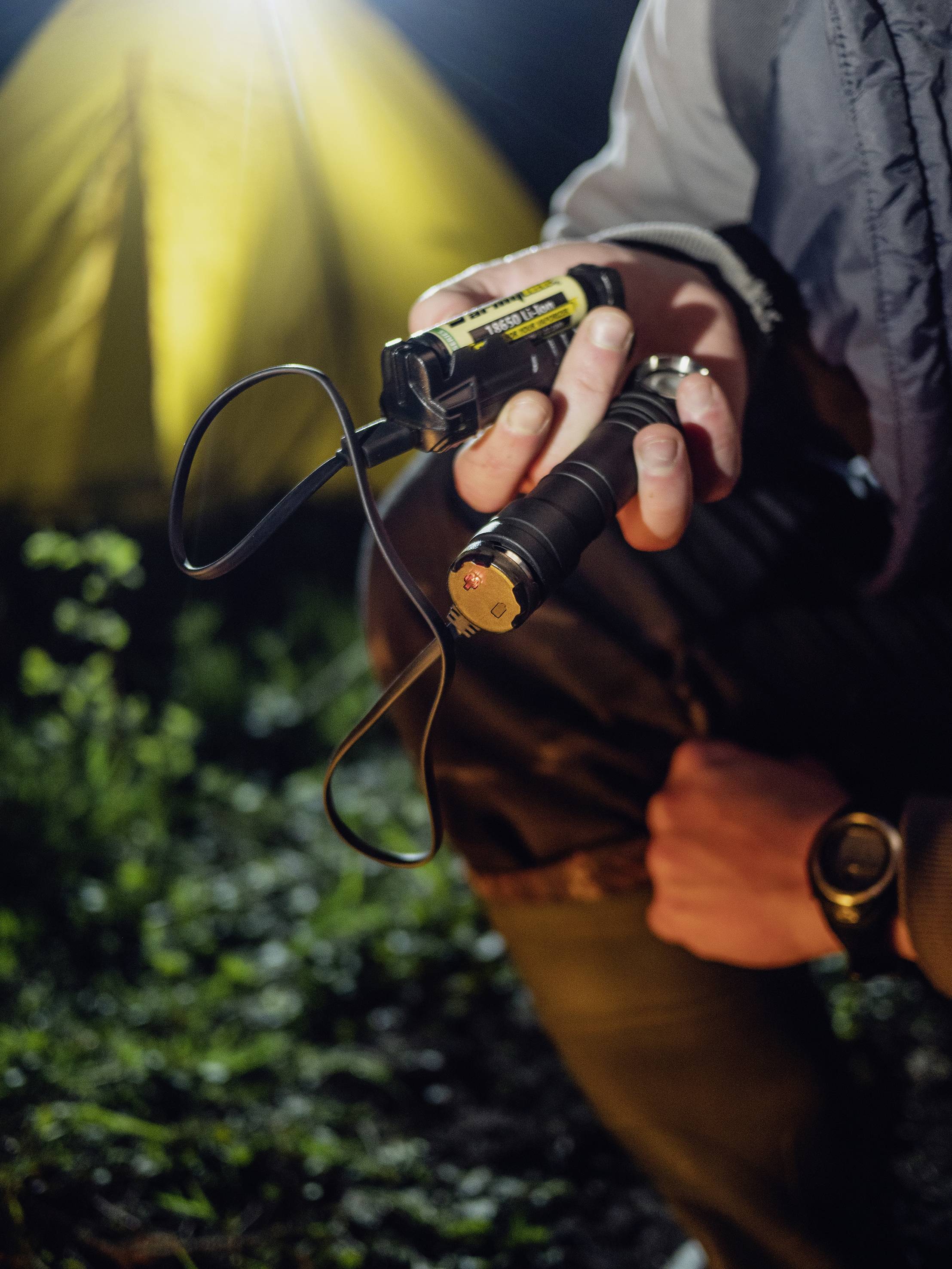 Une personne tient une lampe de poche à la main. À l'arrière-plan, on peut voir une tente illuminée dans la nuit.