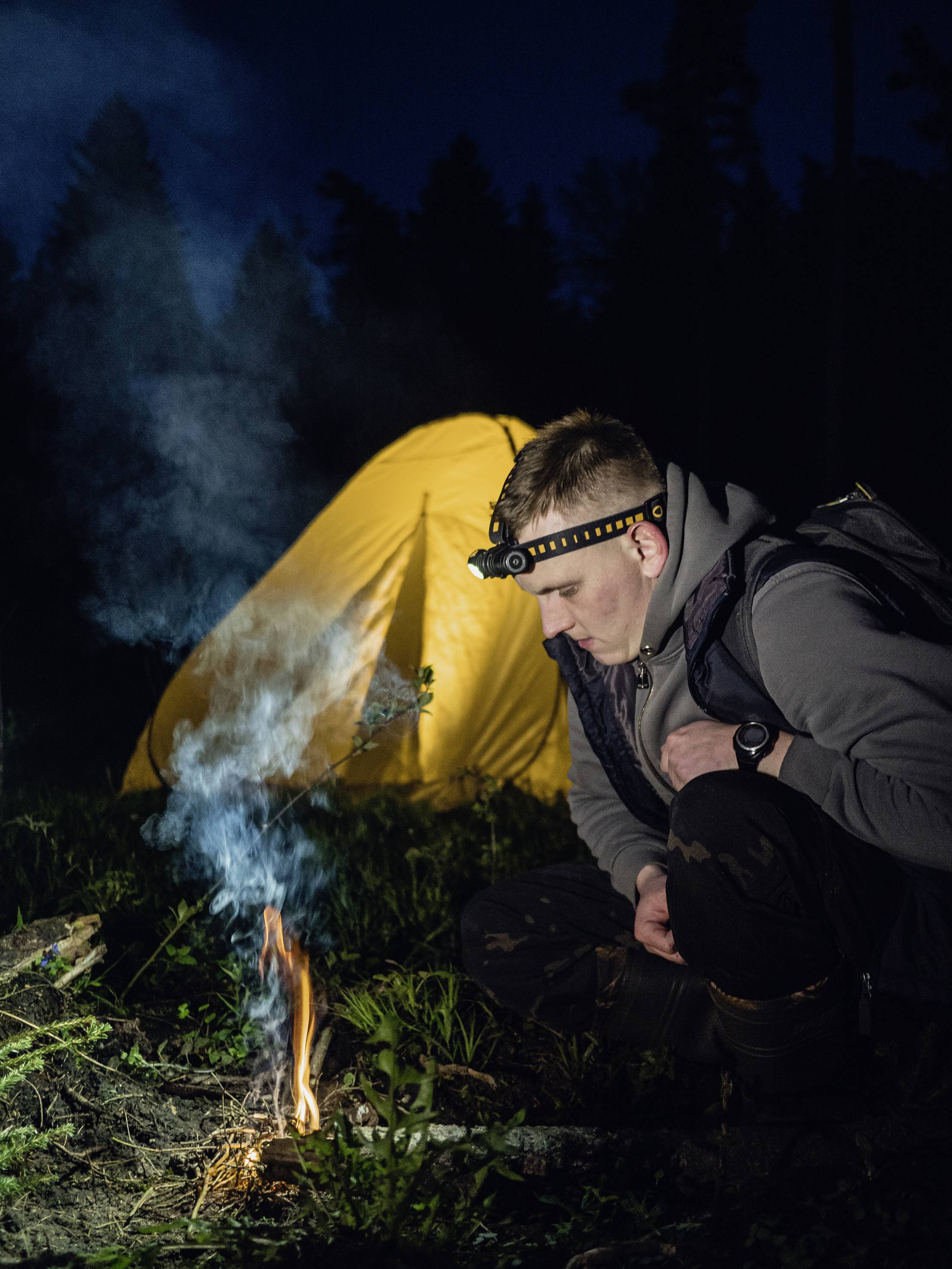 Un homme portant une lampe frontale est accroupi près d'un petit feu de camp la nuit, avec une tente jaune illuminée en arrière-plan.