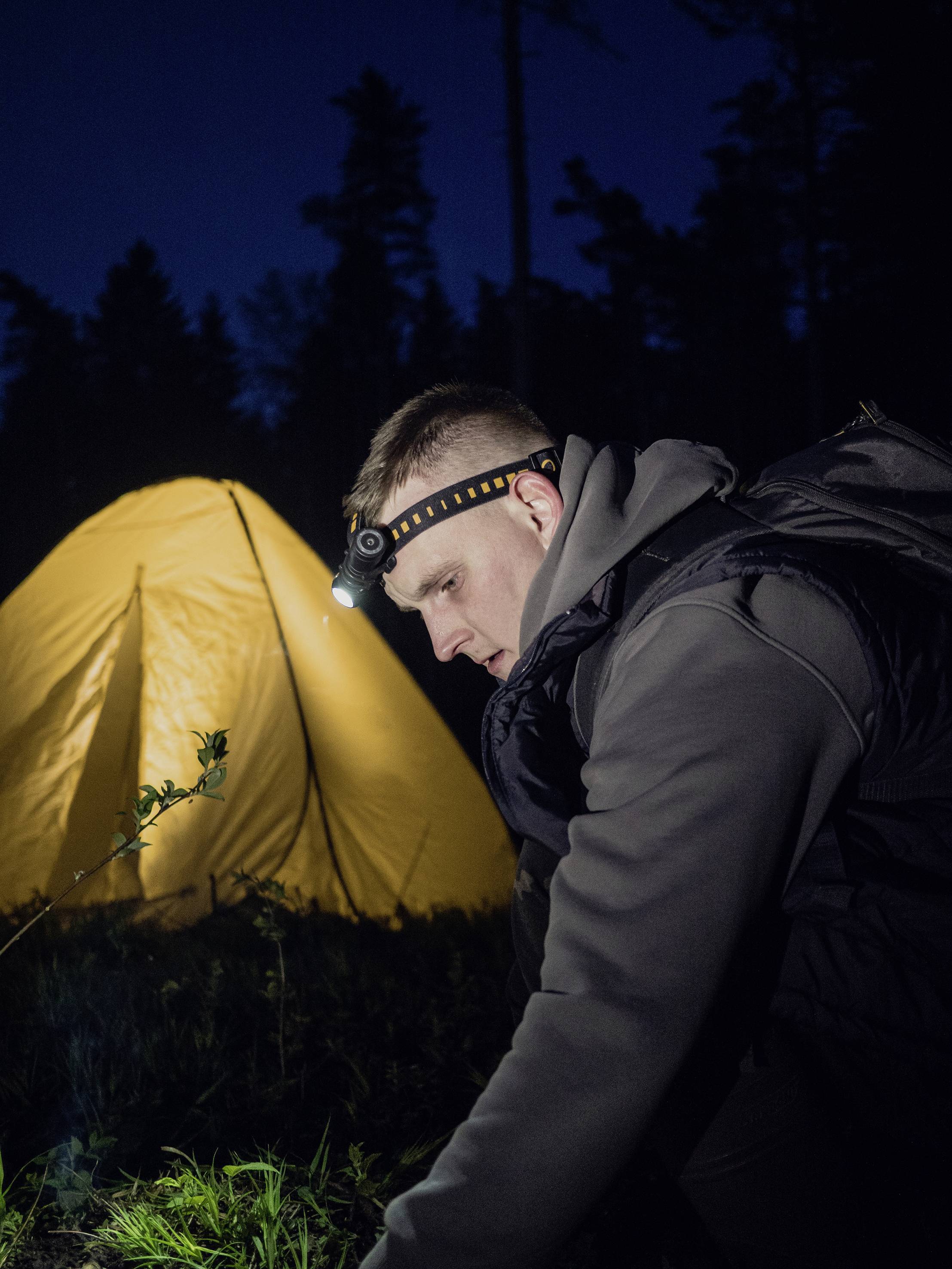 Un homme portant un bandeau frontal est agenouillé dans l'obscurité devant une tente jaune vif dans la forêt et regarde attentivement le sol.