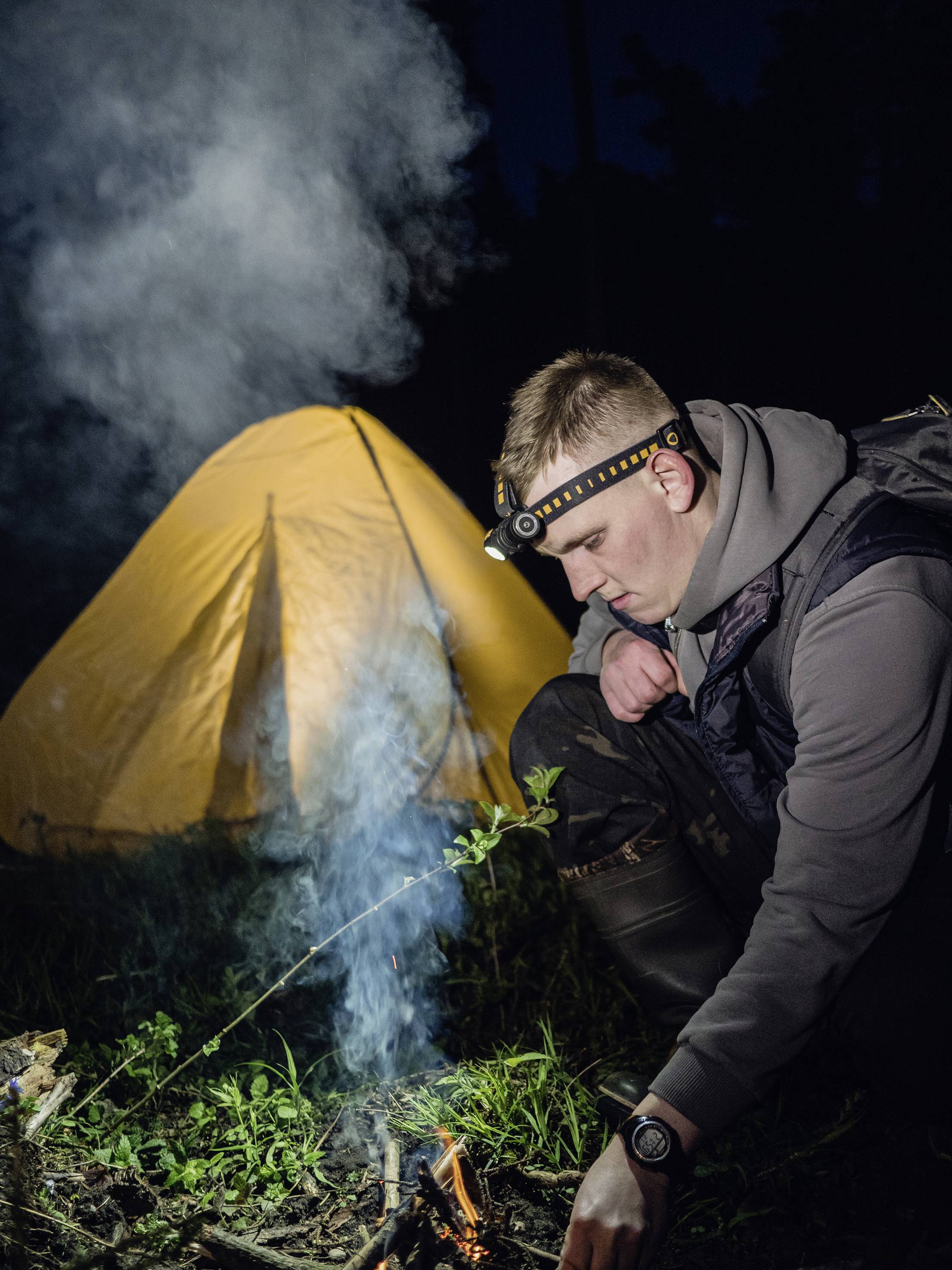 Un homme avec une lampe frontale est agenouillé et allume un feu de camp. À l'arrière-plan, une tente jaune illuminée se dresse dans l'obscurité.
