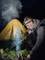 Un homme avec une lampe frontale est agenouillé et allume un feu de camp. À l'arrière-plan, une tente jaune illuminée se dresse dans l'obscurité.