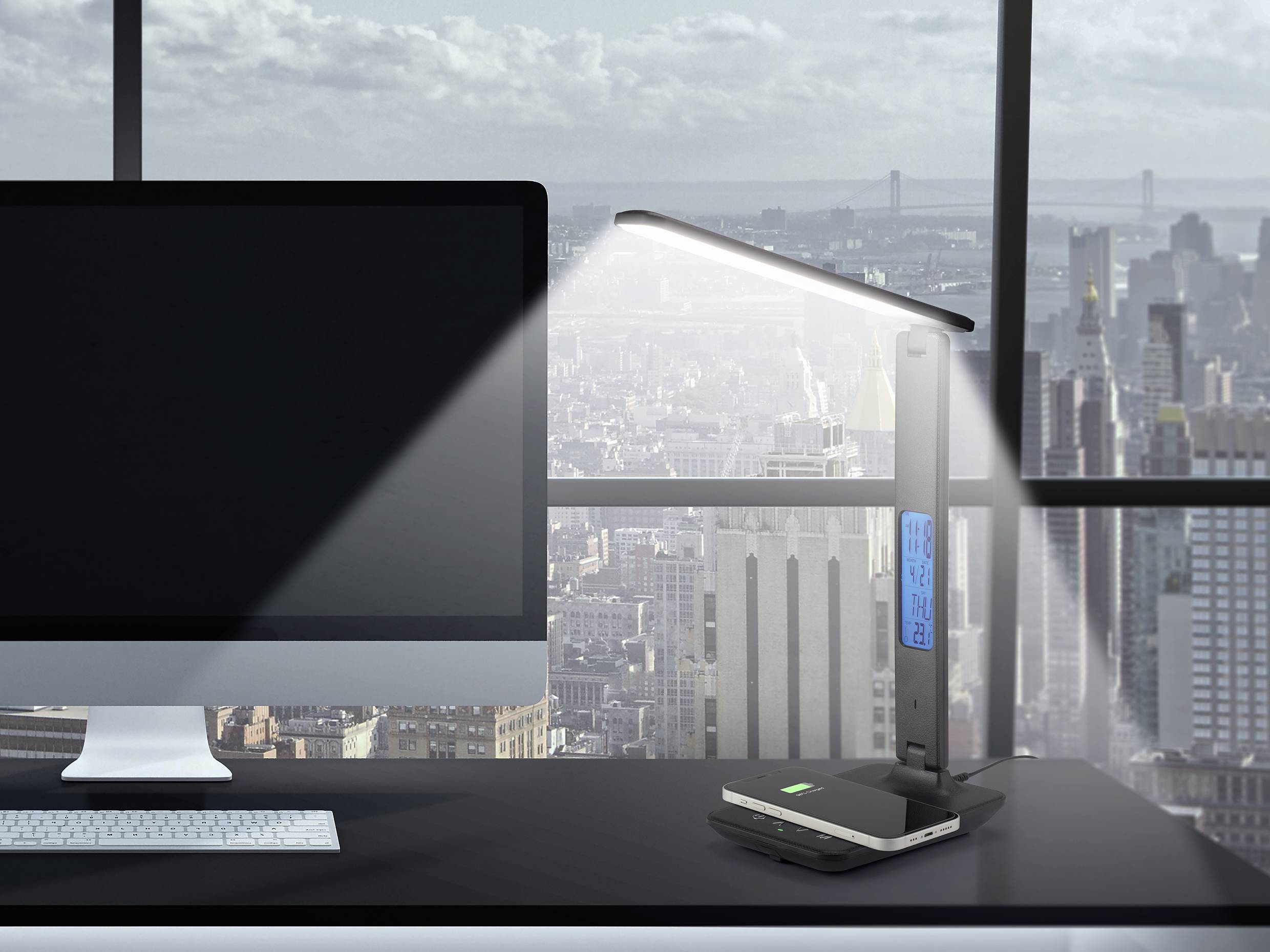 Bureau avec un ordinateur et une lampe de bureau éclairant un smartphone. En arrière-plan : une fenêtre donnant sur un paysage urbain.