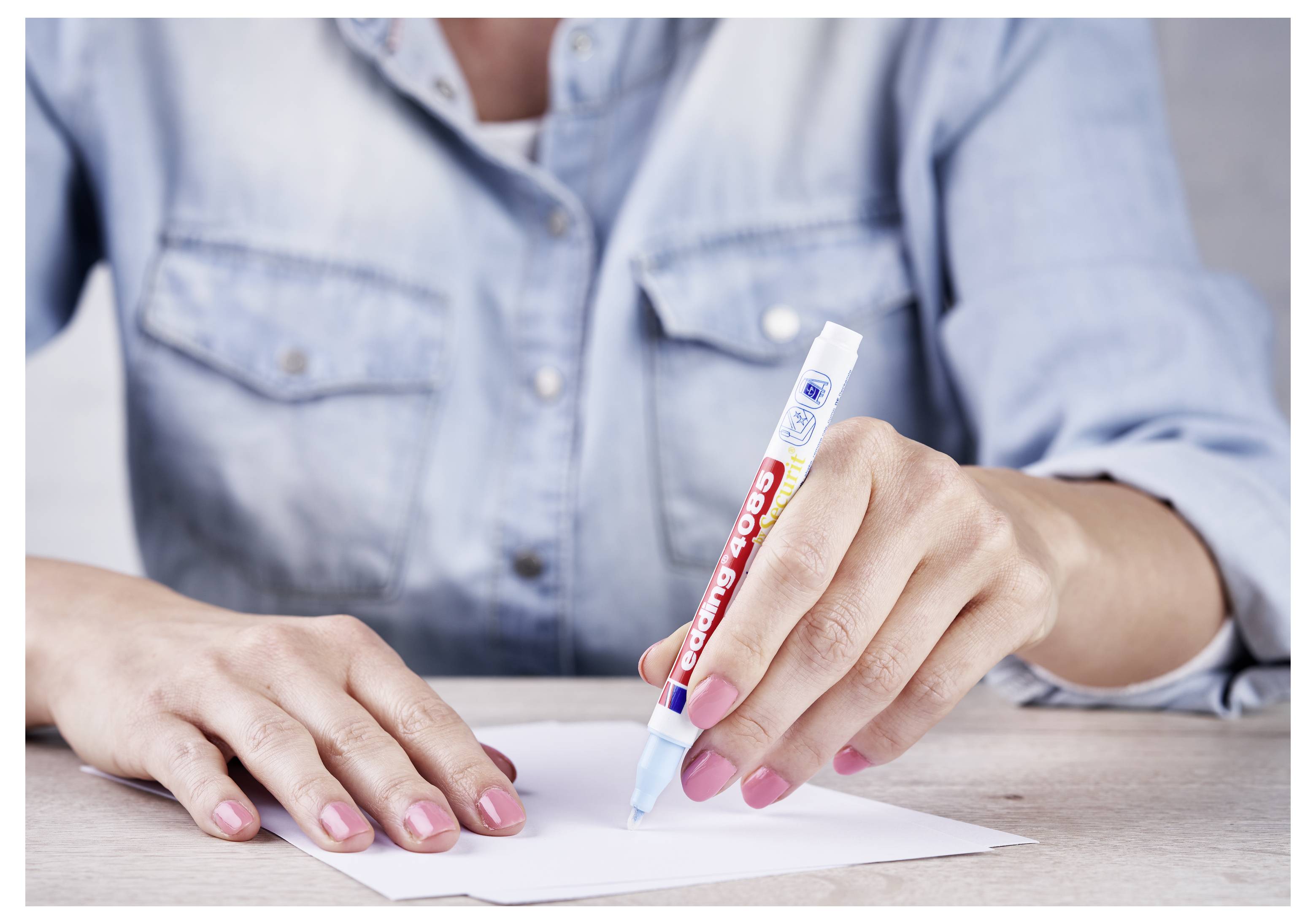 Une personne vêtue d'une chemise en denim écrivant sur une feuille de papier avec un stylo bleu et rouge à un bureau.