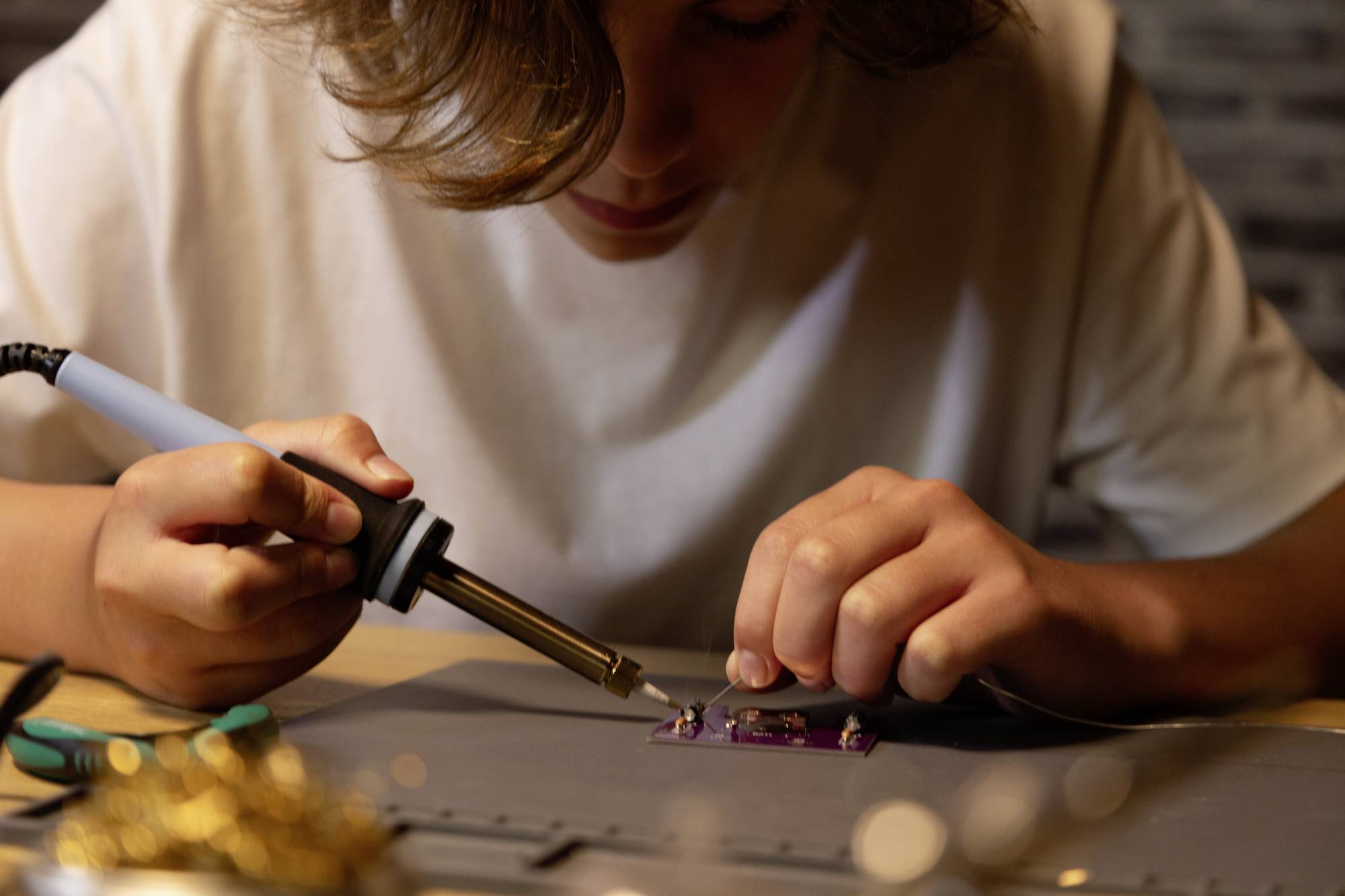 Une personne soude attentivement un petit circuit électronique. Des outils sont visibles à côté du fer à souder.