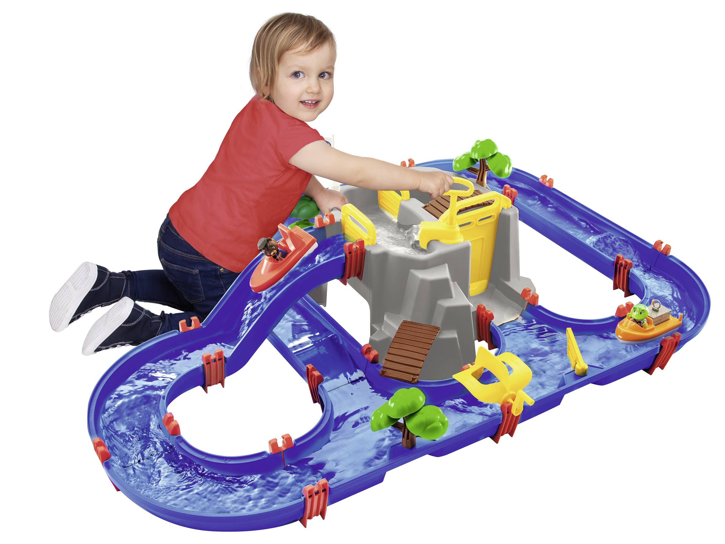 Un enfant sourit en jouant avec un circuit aquatique bleu, comprenant des bateaux et des écluses. Il porte un t-shirt rouge et est assis par terre.