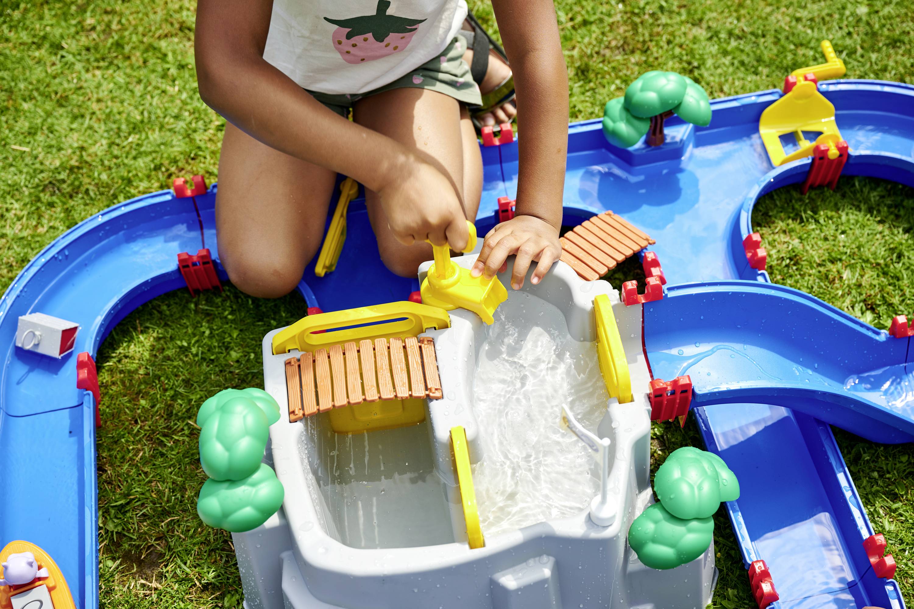 Un enfant joue avec un jouet aquatique bleu, doté de canaux et de systèmes d'écluses, à l'extérieur sur une pelouse.