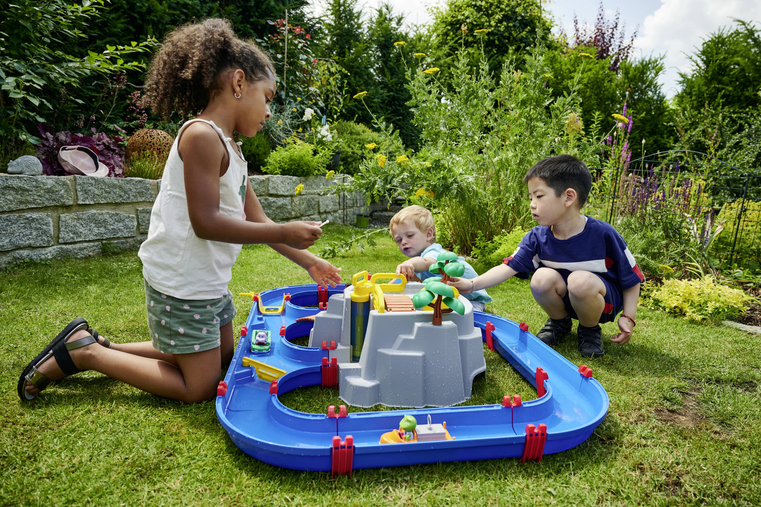 Trois enfants jouent dans le jardin avec un train miniature qui circule autour d'une structure centrale grise. Ils sont dans un environnement verdoyant.