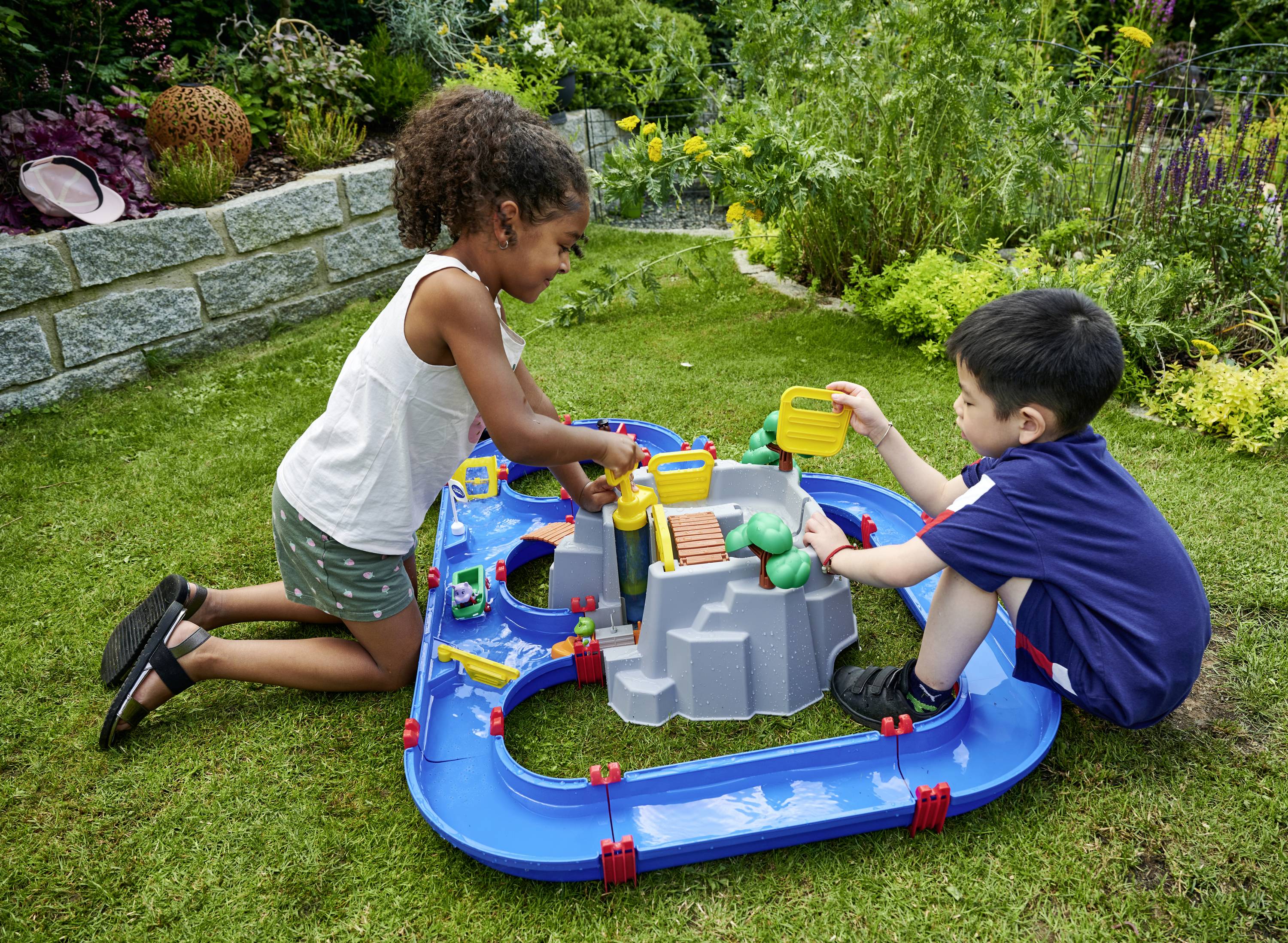 Deux enfants jouent dans le jardin avec un toboggan aquatique bleu, puisant de l'eau avec des arrosoirs jaunes. Des plantes sont visibles en arrière-plan.