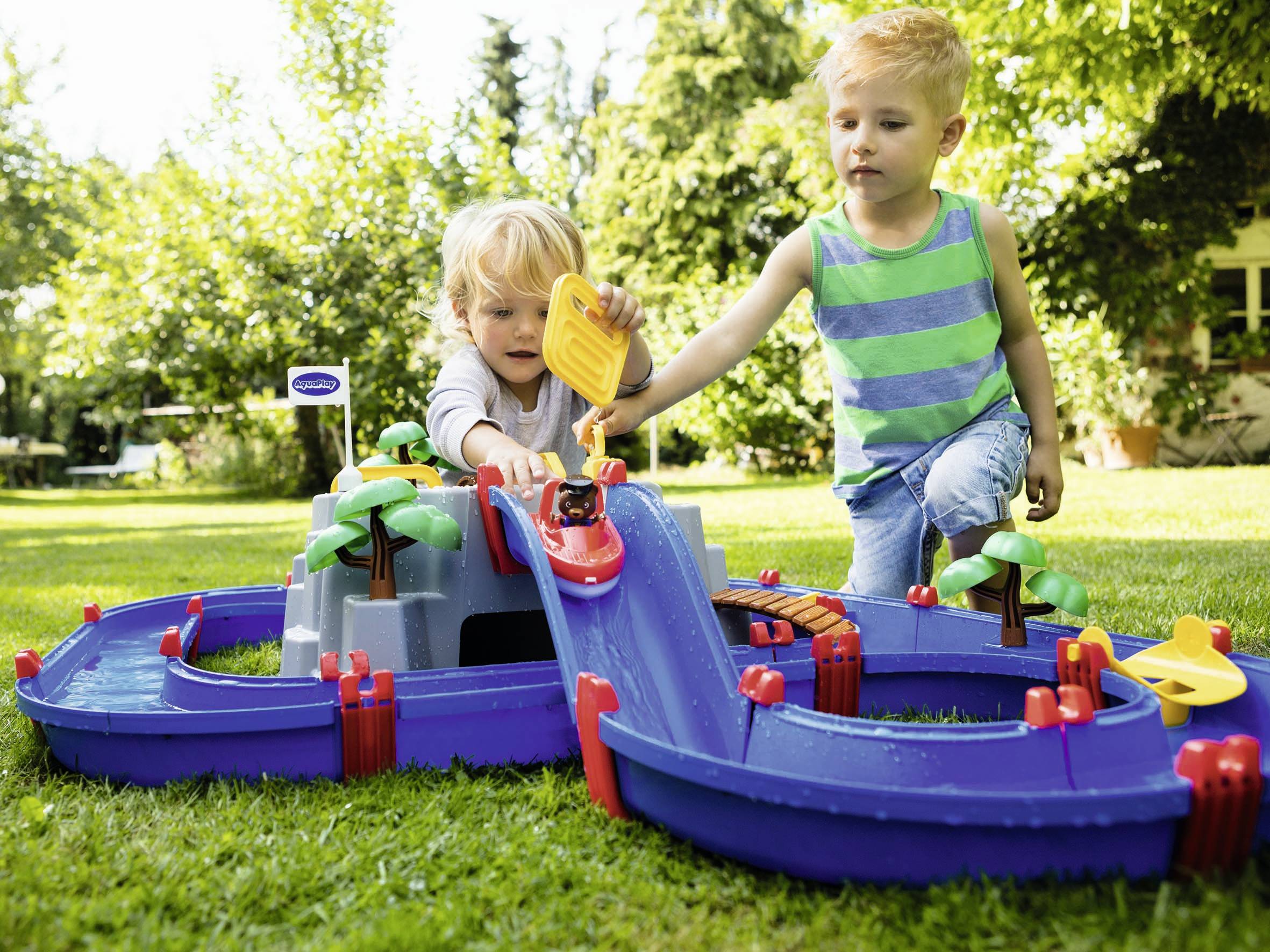 Deux enfants jouent à l'extérieur avec un ensemble de circuit aquatique bleu. Ils versent de l'eau dedans, tandis que des arbres en plastique entourent le canal.