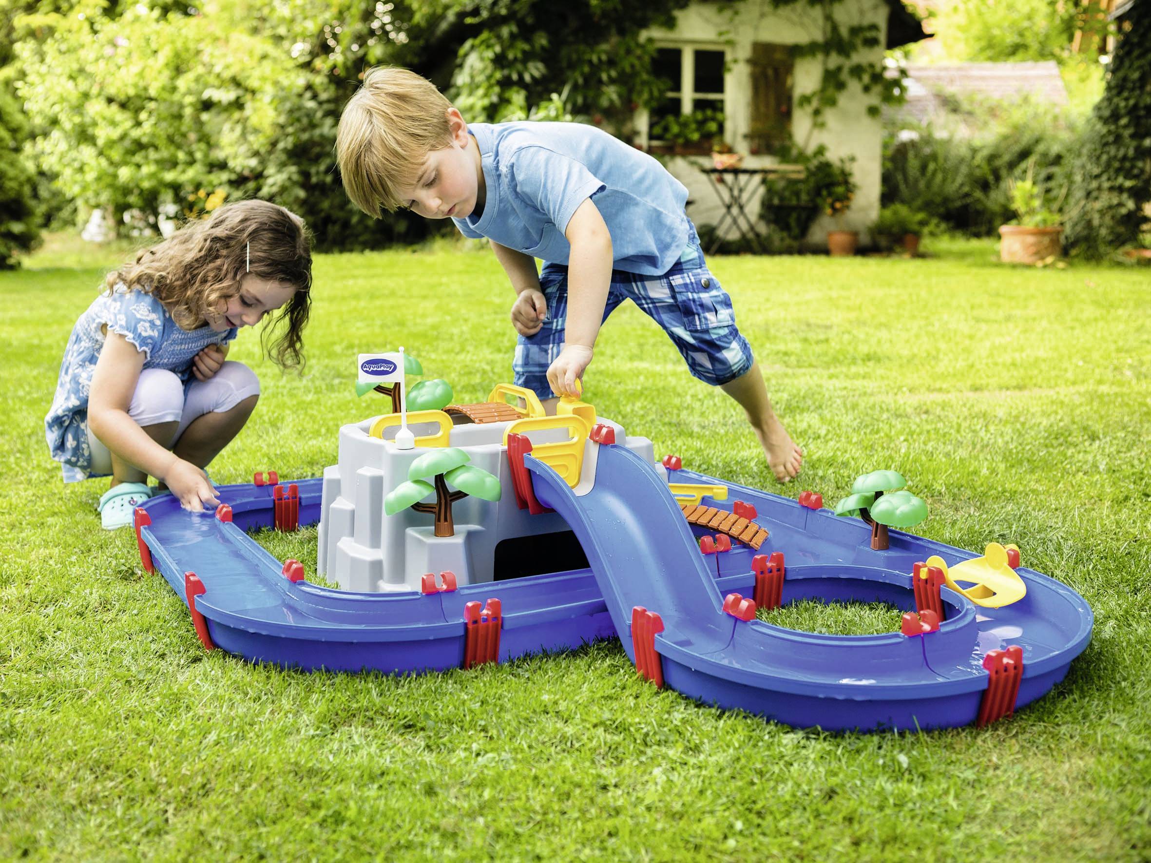 Deux enfants jouent dans le jardin avec un toboggan aquatique. L'ensemble comprend des canaux, des ponts et de petits bateaux qui flottent sur l'eau.