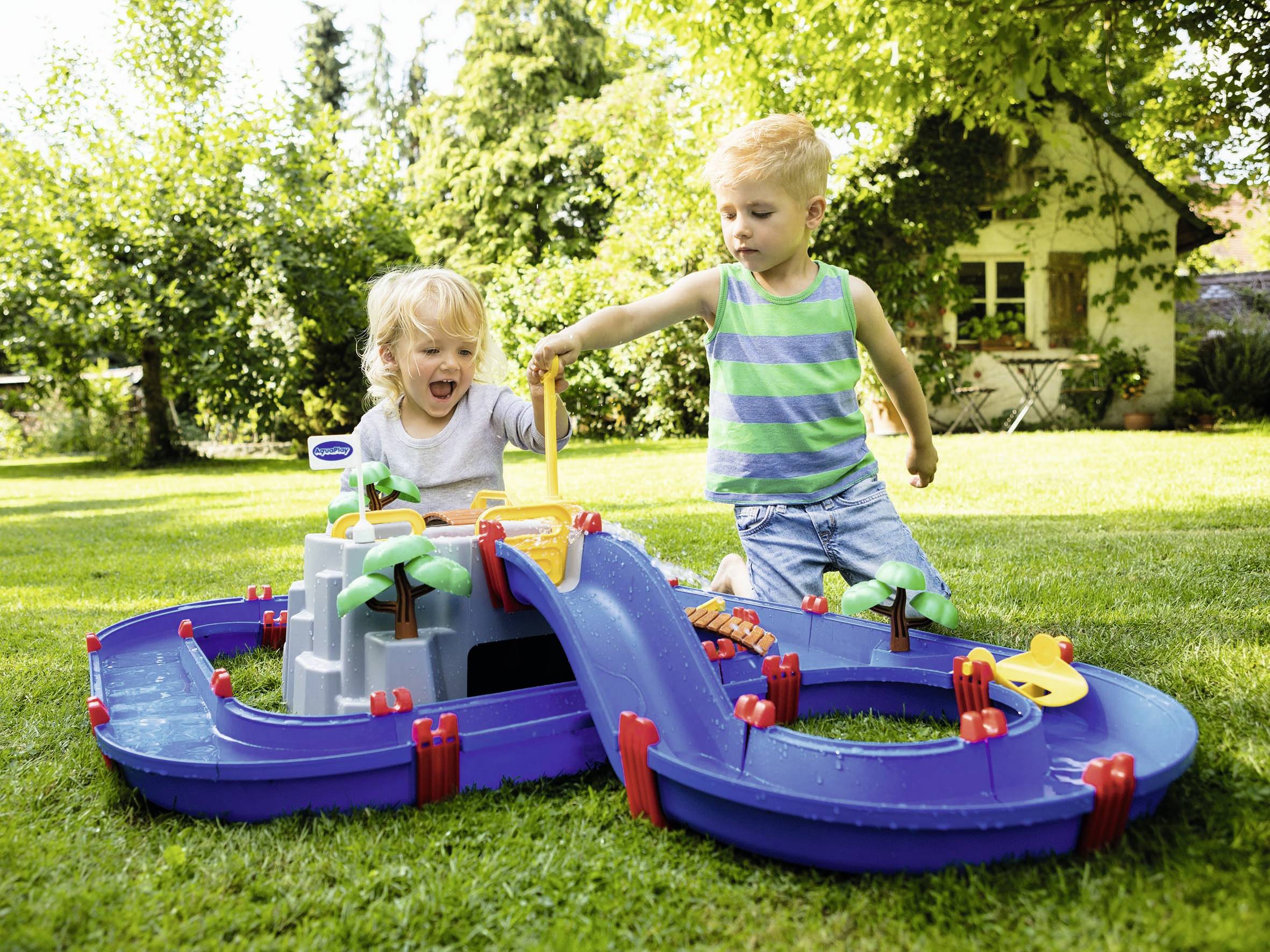 Deux enfants jouent dans le jardin avec un toboggan aquatique bleu. Un garçon porte un t-shirt rayé, l'autre enfant se réjouit du jeu. À l'arrière-plan, une maison est visible.