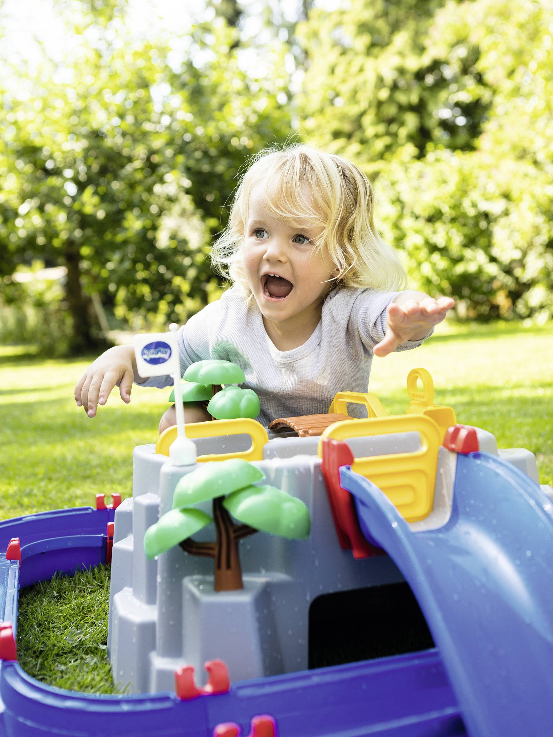 Un petit enfant joue dehors avec un château de jouet coloré avec un toboggan et des arbres, entouré d'herbe verte et d'arbres en arrière-plan.
