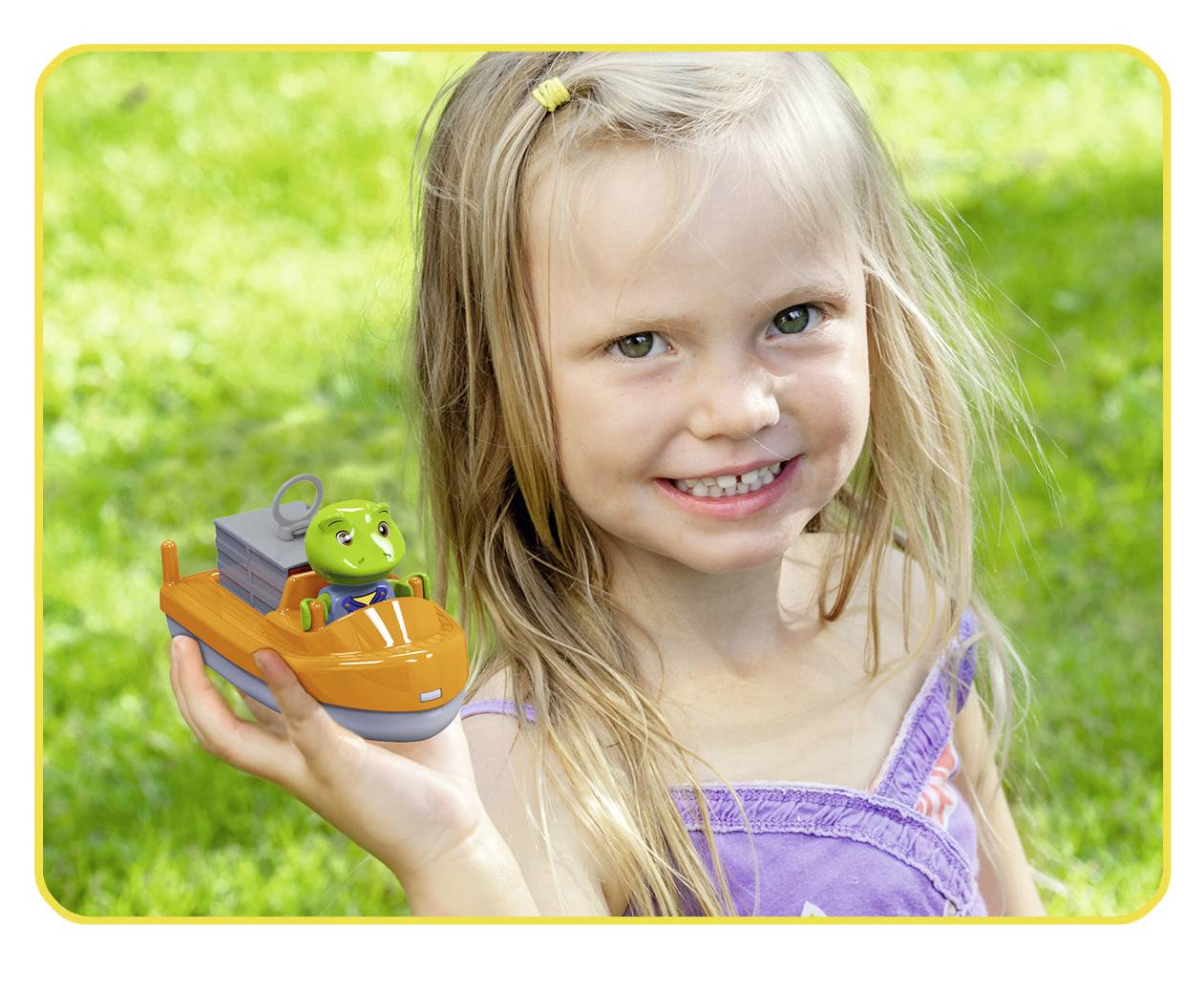 Une petite fille tient un petit bateau jouet orange avec une figurine verte dessus, sourit dans un jardin ensoleillé.