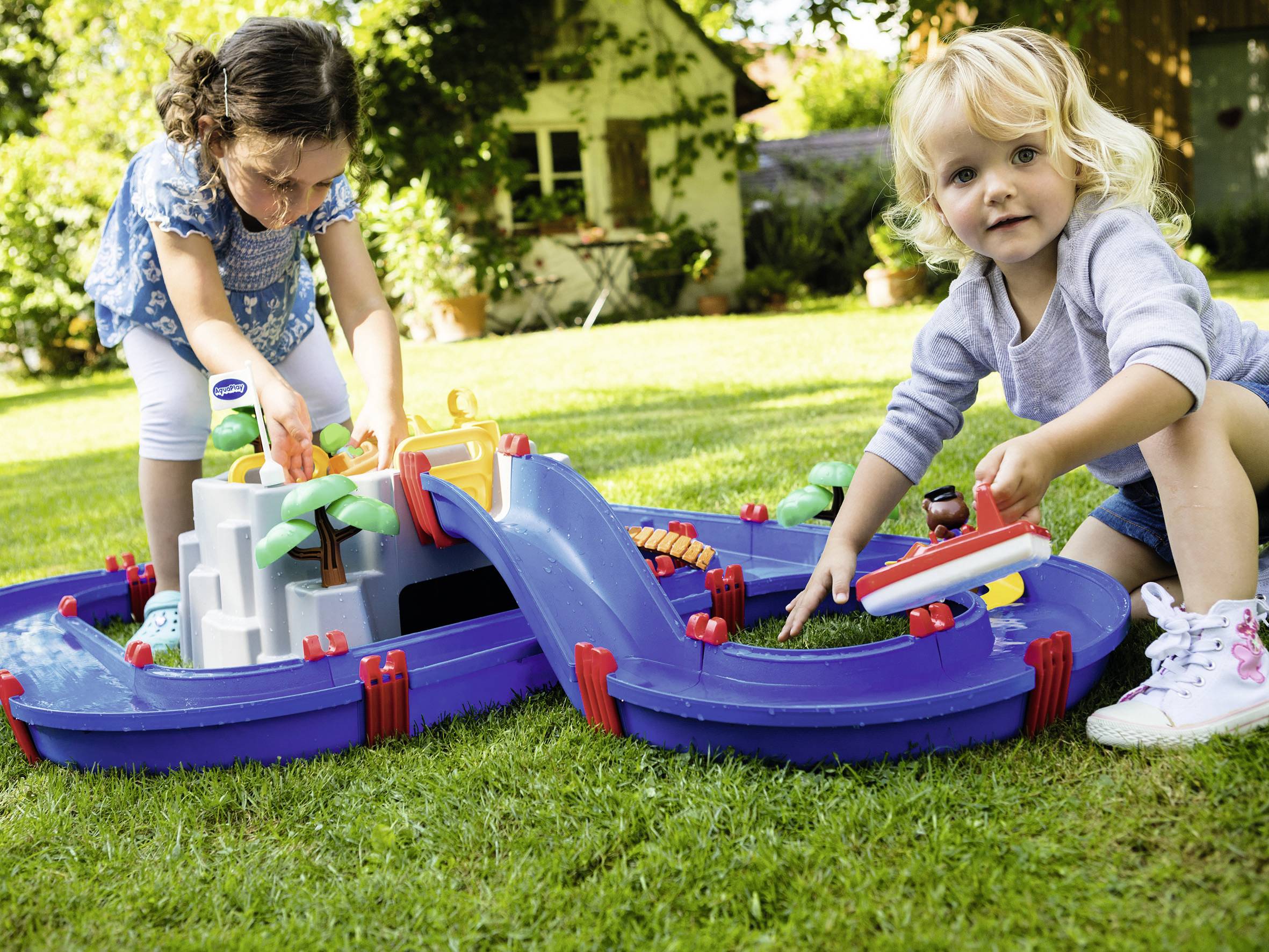 Deux enfants jouent dans le jardin avec un toboggan aquatique bleu, qui contient des palmiers et des bateaux. Une maison est visible en arrière-plan.