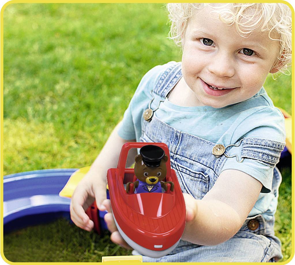 Un enfant aux boucles blondes tient un bateau jouet rouge avec une figurine portant un costume et un chapeau haut-de-forme. De l'herbe est visible en arrière-plan.