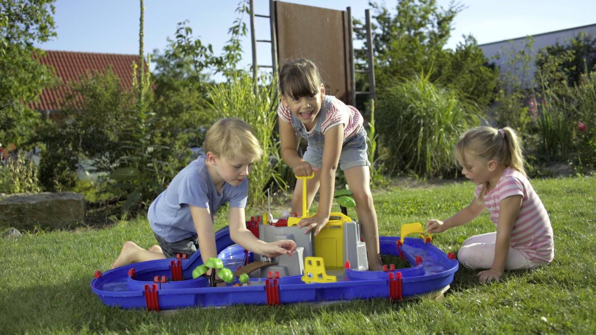 Trois enfants jouent dehors sur une prairie avec un toboggan aquatique bleu. En arrière-plan, on peut voir des arbres et des bâtiments.