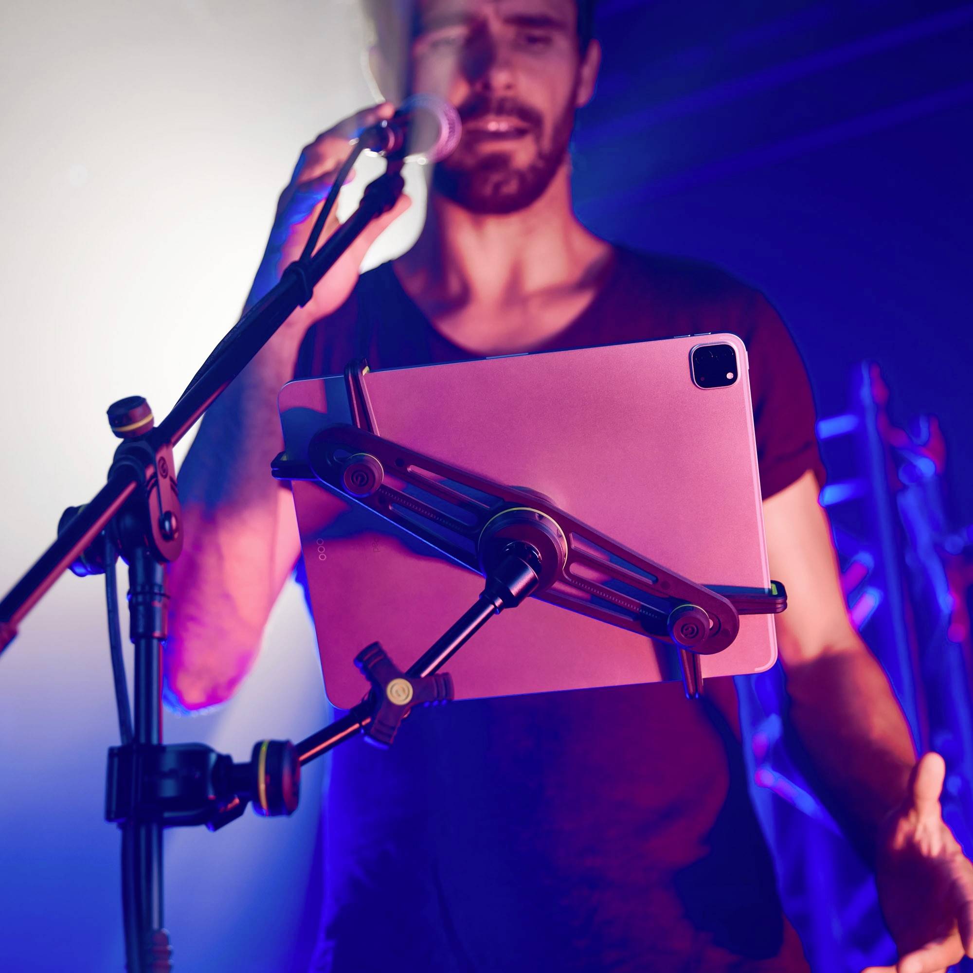Un homme chante dans un microphone tout en regardant une tablette fixée sur un support. Scène en arrière-plan.