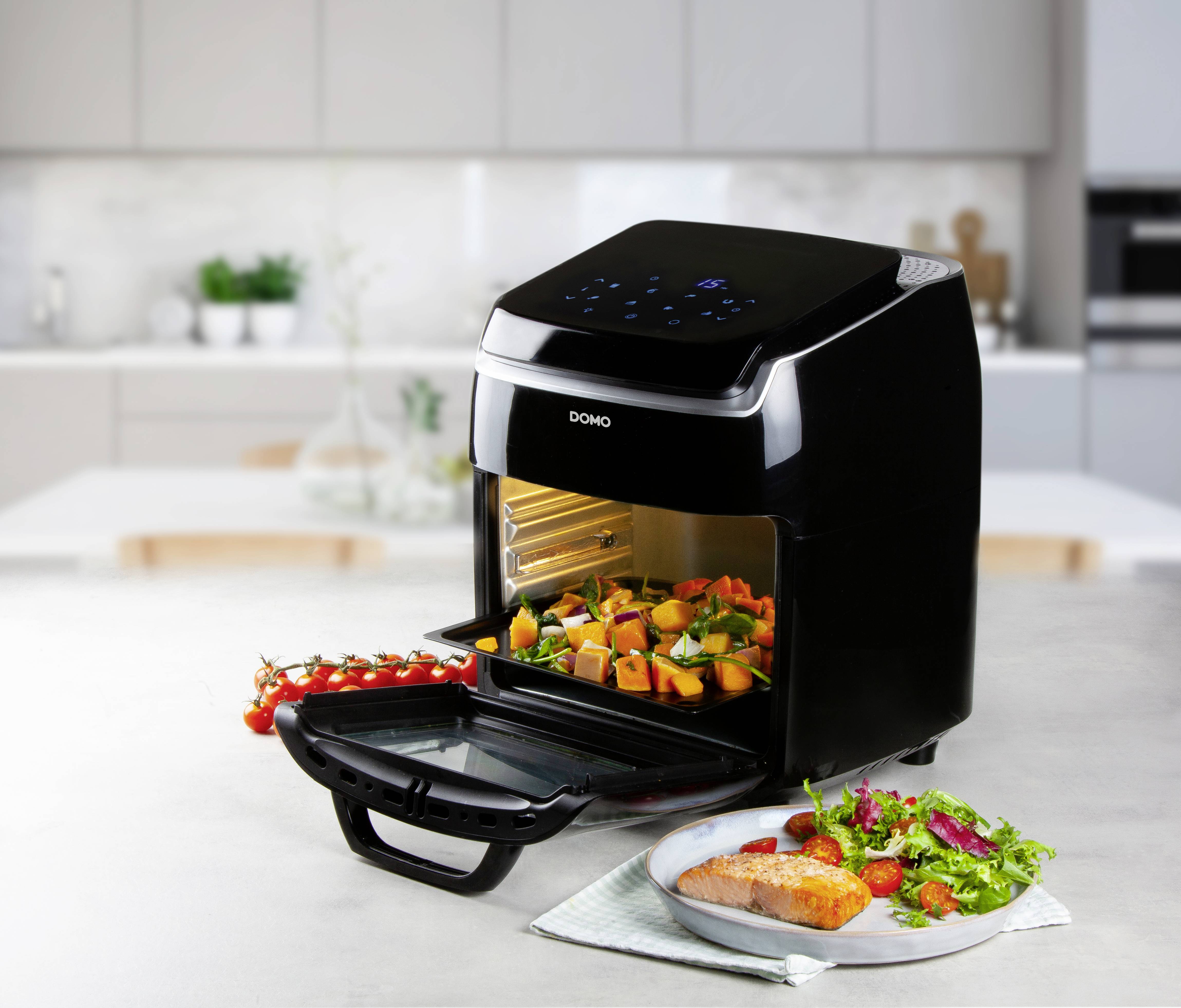 Un four à convection noir est posé sur un comptoir de cuisine. La porte est ouverte, et des légumes sont à l'intérieur. À côté, il y a une assiette avec du saumon et de la salade.