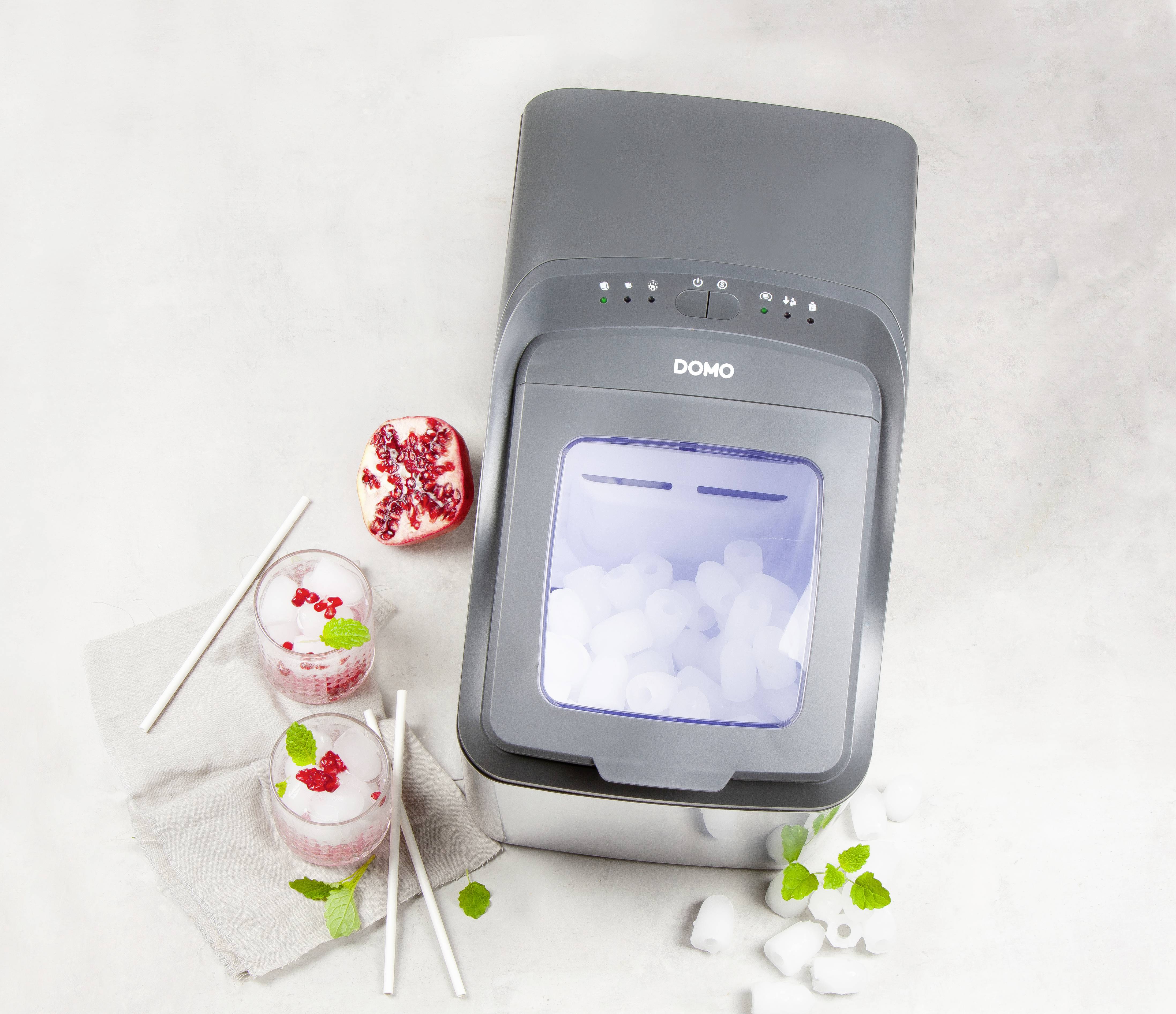 Une machine à glaçons compacte avec des glaçons dans le récipient, entourée de deux verres de boissons, de glaçons, de graines de grenade et de feuilles de menthe.