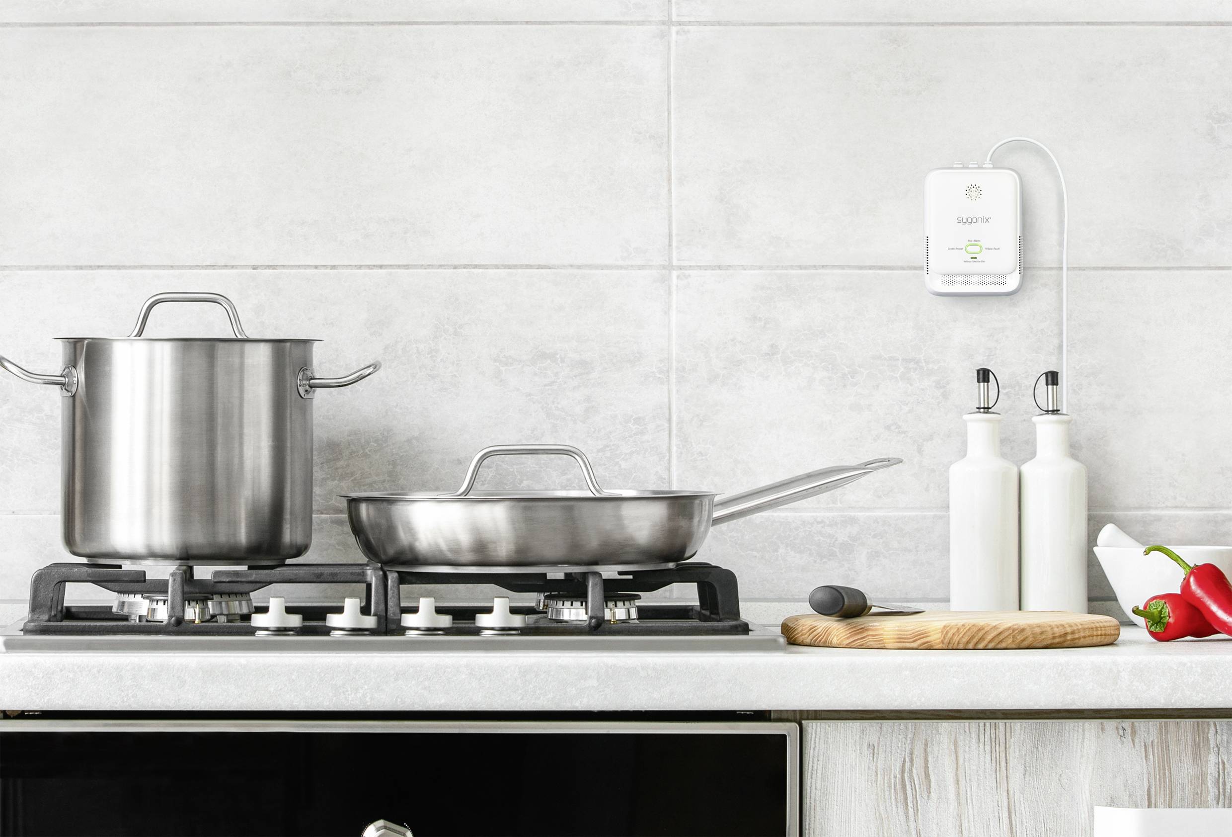 Cuisinière avec casserole et poêle, à côté des bouteilles d'huile et une planche à découper avec des poivrons. Un capteur blanc est accroché au mur.