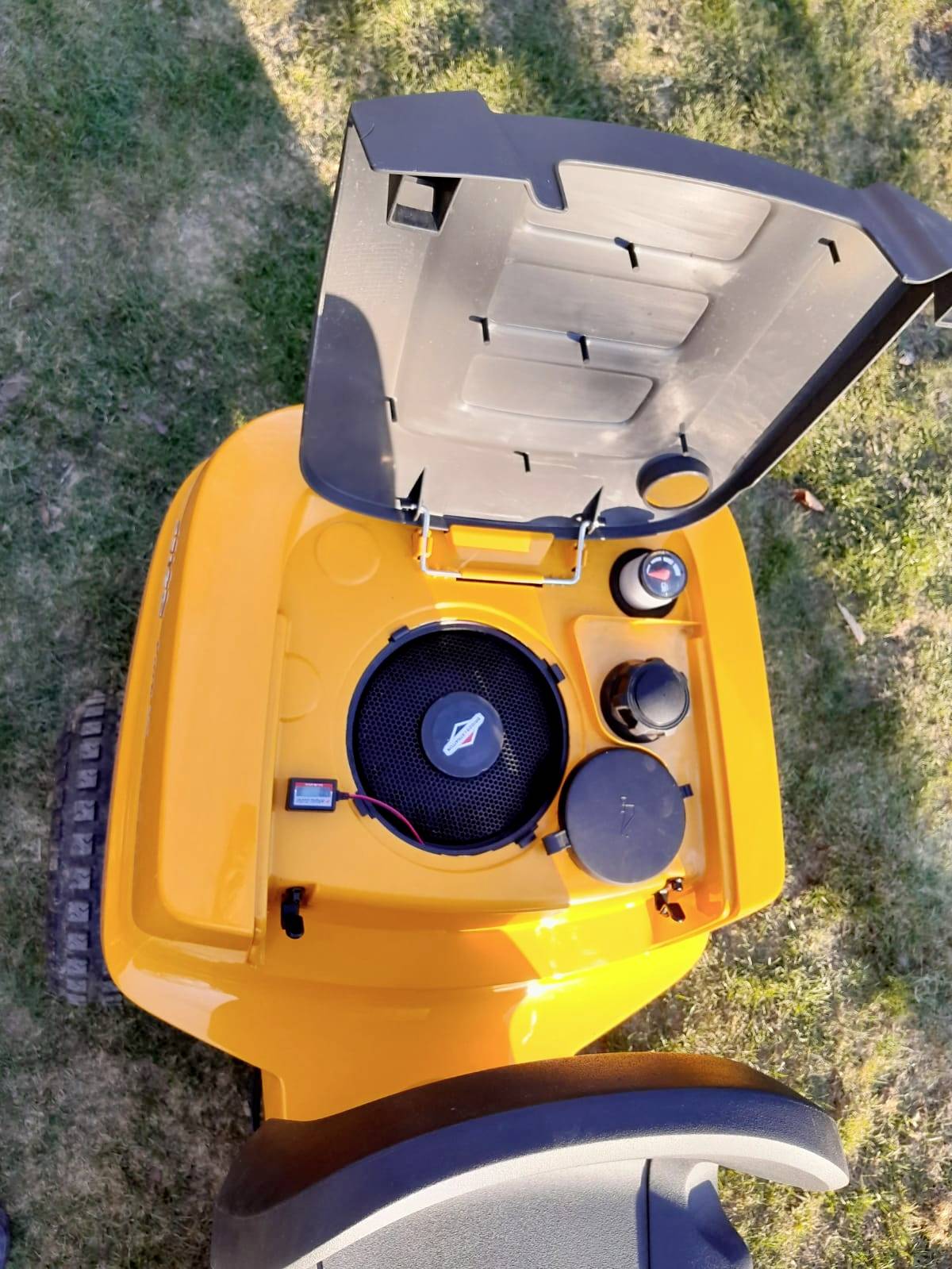 Tondeuse à gazon jaune vue du dessus, capot moteur ouvert. Le moteur est visible, et la tondeuse est posée sur de l'herbe verte.