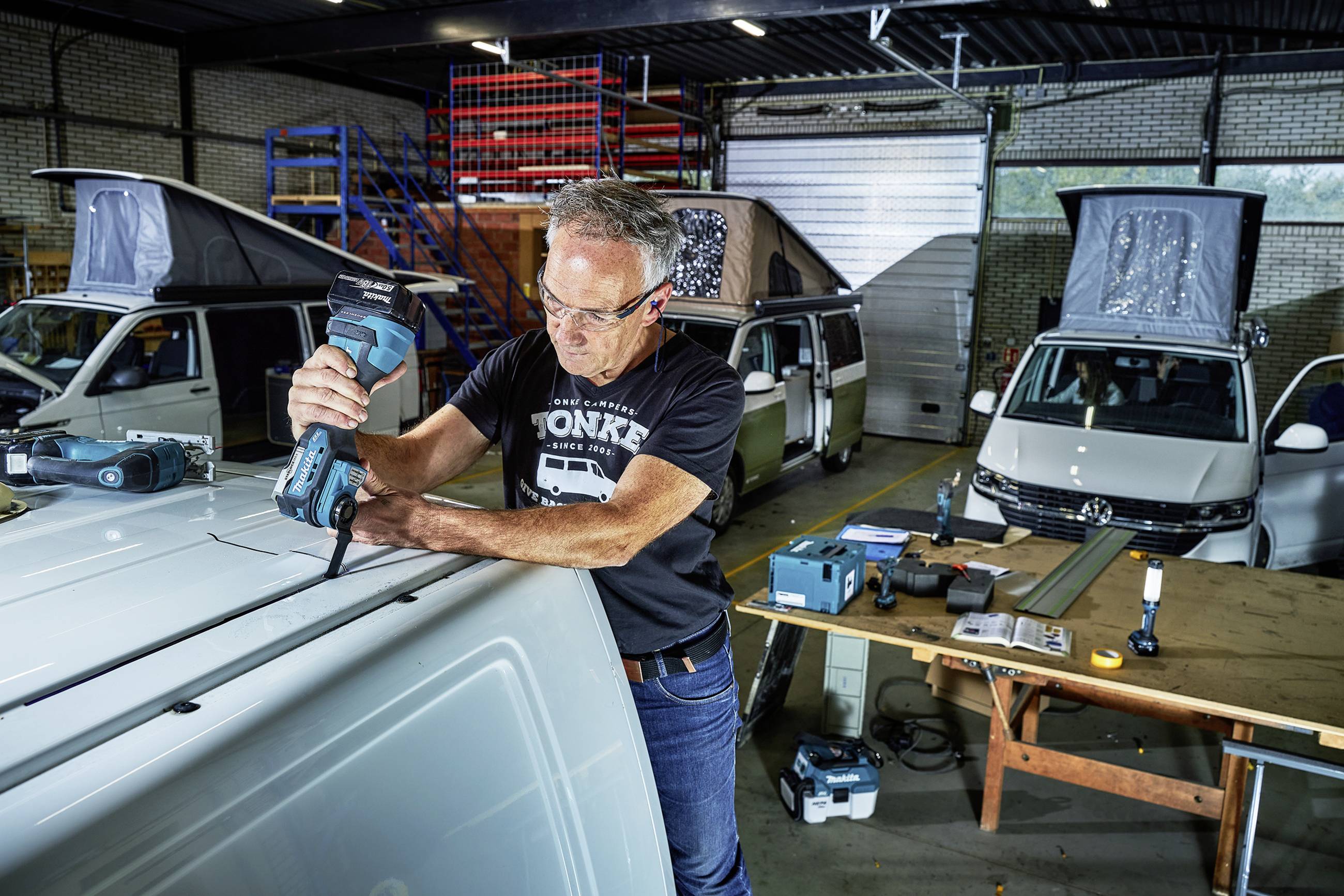 Un homme travaille sur un véhicule blanc avec un outil électrique dans un atelier. D'autres véhicules sont visibles en arrière-plan.