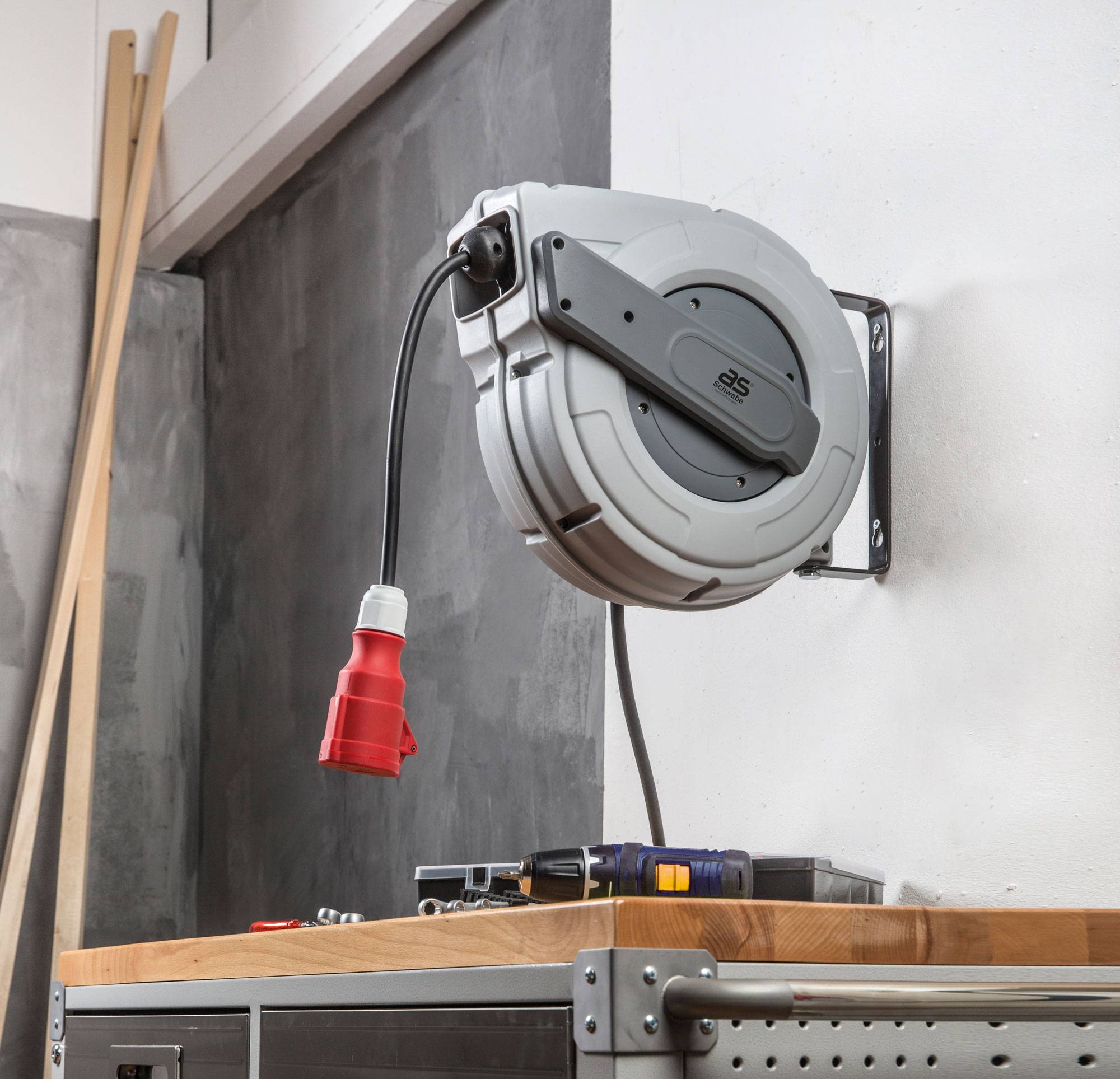 Enrouleur de câble automatique fixé au mur, avec câble électrique rétractable suspendu. Outils posés sur la table au premier plan.