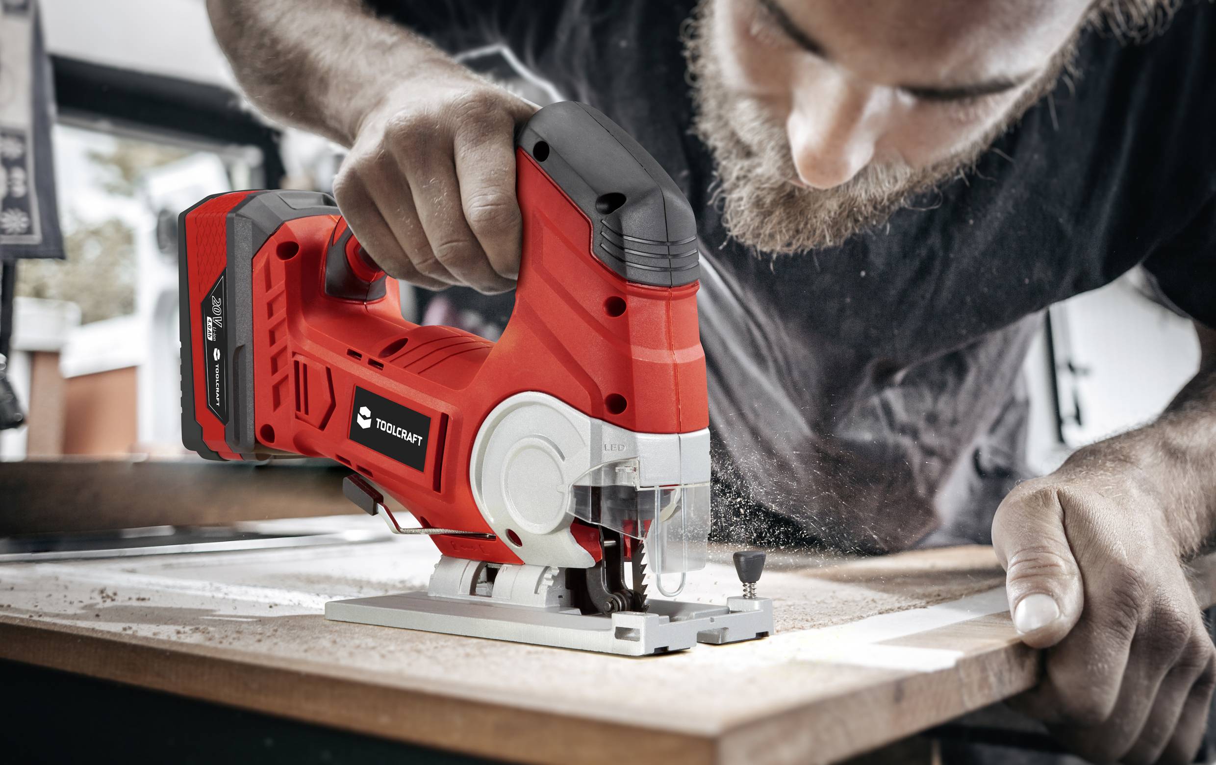 Un homme utilise une scie sauteuse rouge pour couper une planche. De la poussière voltige dans l'air. Expression concentrée. Gros plan.
