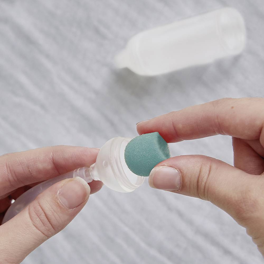 Une main tient un bouchon plat et rond ; une autre main le colore avec une éponge turquoise.