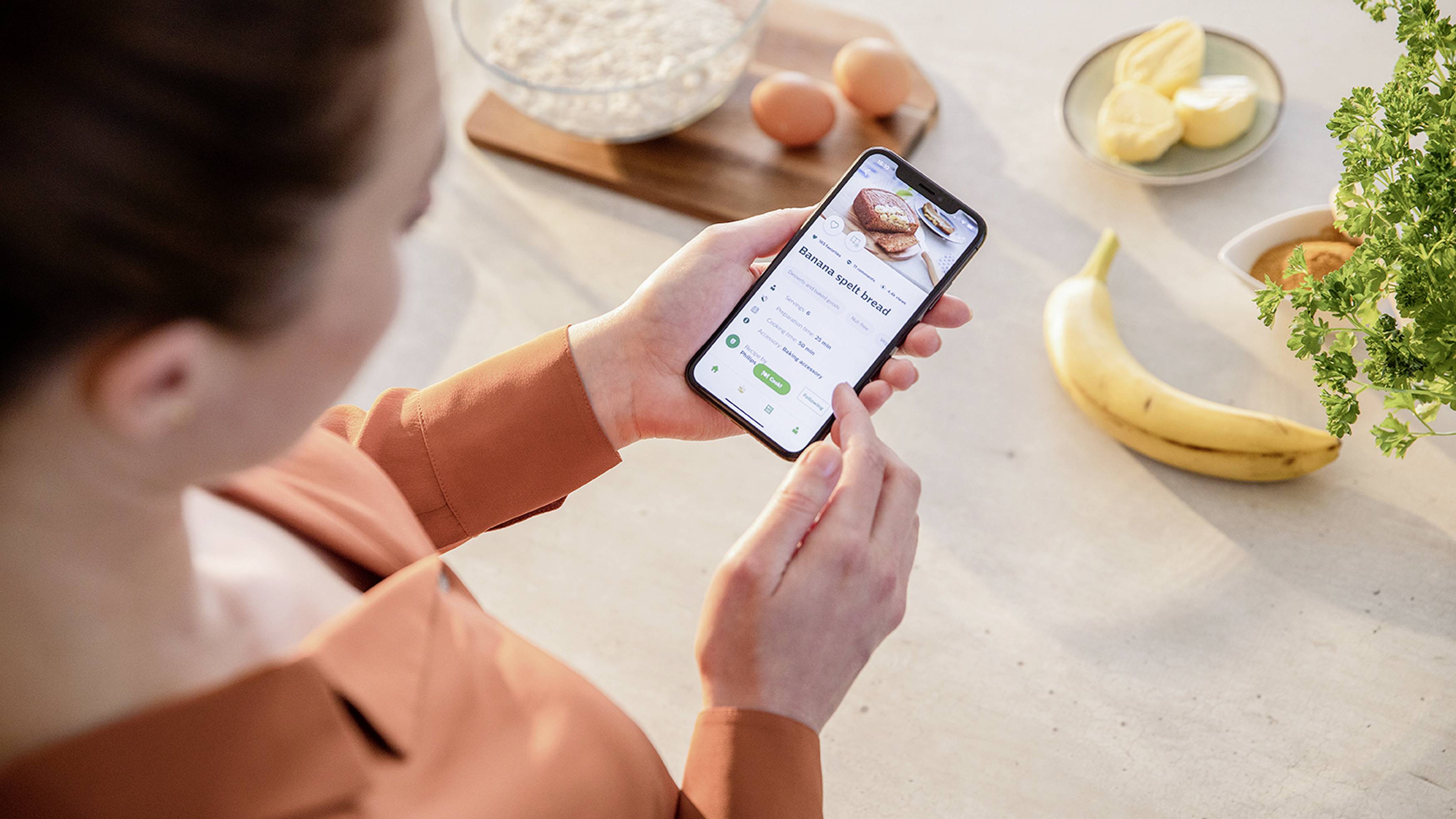 Une femme dans la cuisine tient un smartphone avec une application de recettes de cuisine, à côté d'un bol, d'œufs, de beurre, d'une banane et de légumes verts sur la table.
