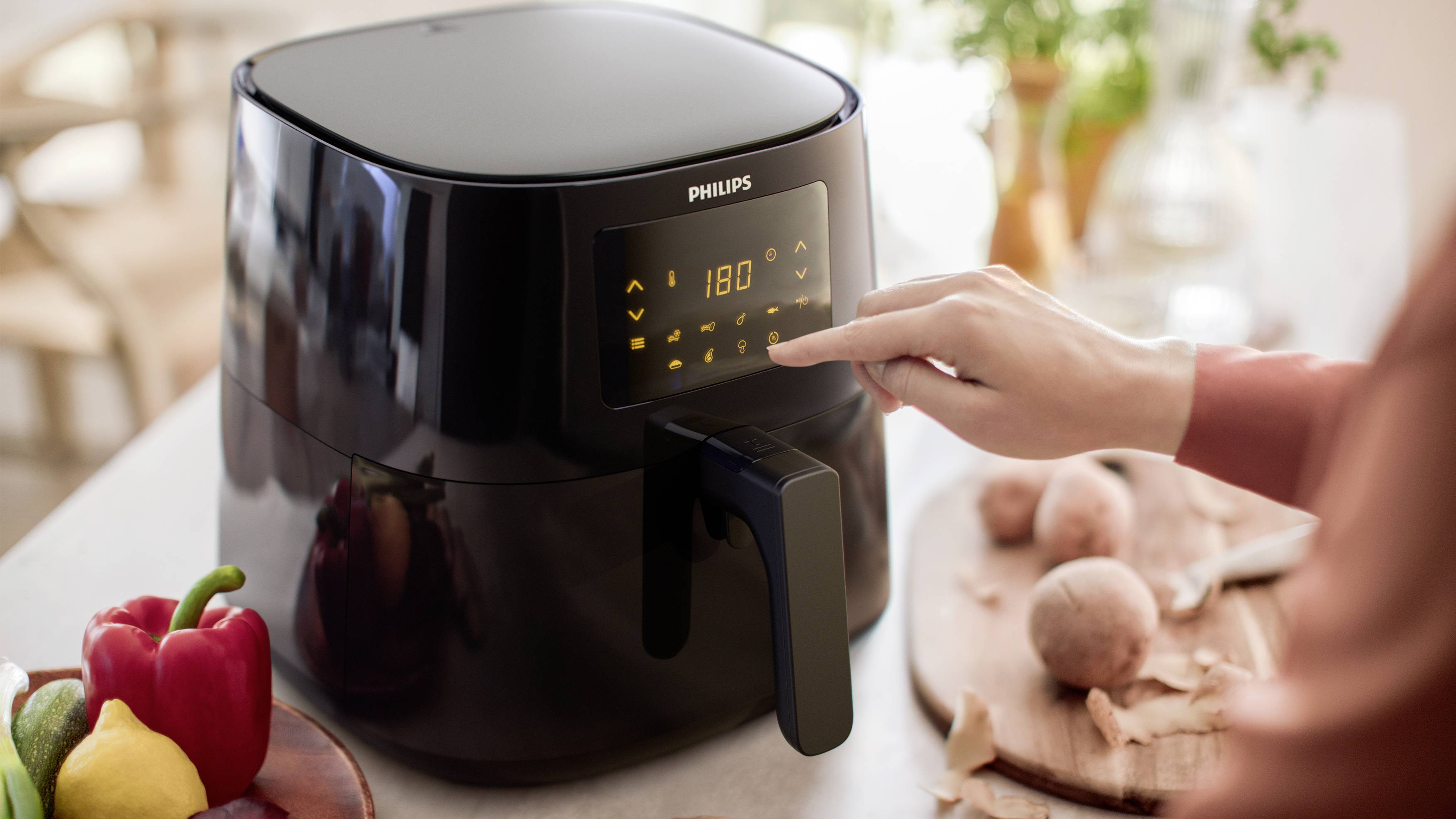 Une personne utilise une friteuse à air chaud noire sur un plan de travail de cuisine. Des poivrons et des pommes de terre coupées sont disposés à côté.